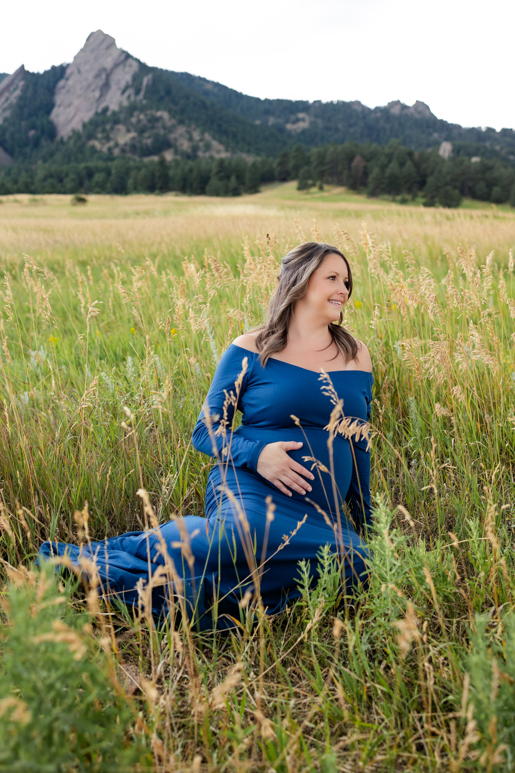 Pregnant woman in a blue dress sits in a mountain field with one hand on her belly smiling off camera.