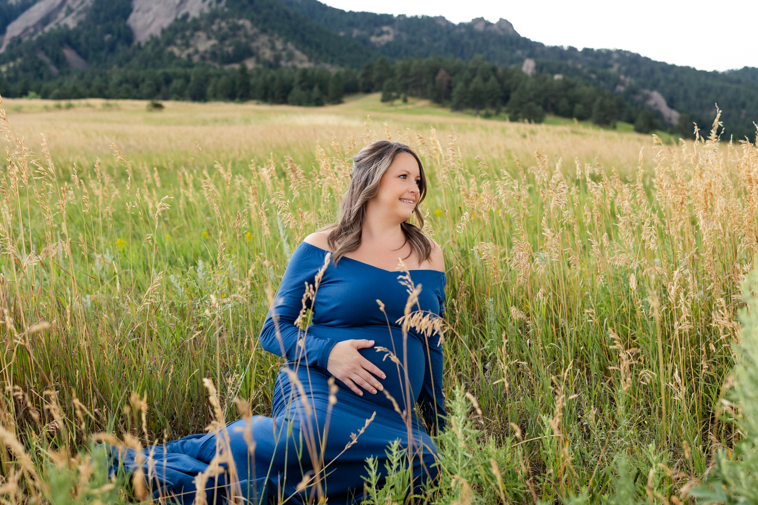 Pregnant woman in a blue dress sits in a mountain field with one hand on her belly smiling off camera.