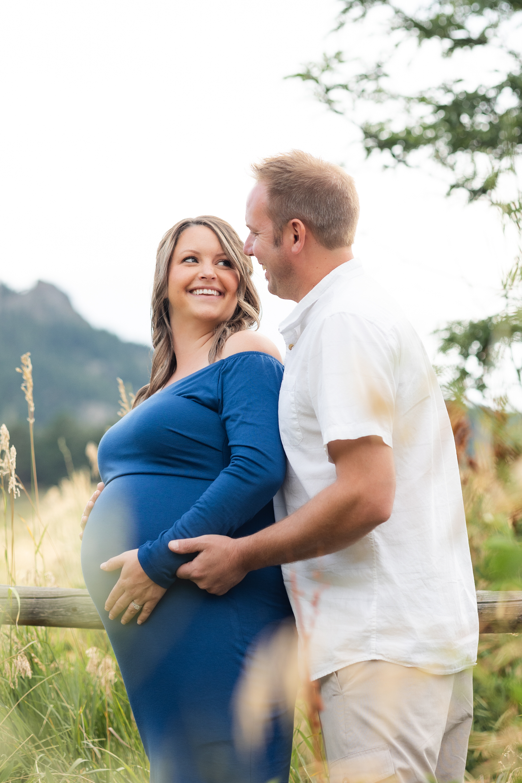 A pregnant woman stands with her back to her husband's chest and smiles over her shoulder at him while they both have their hands on her pregnant belly.