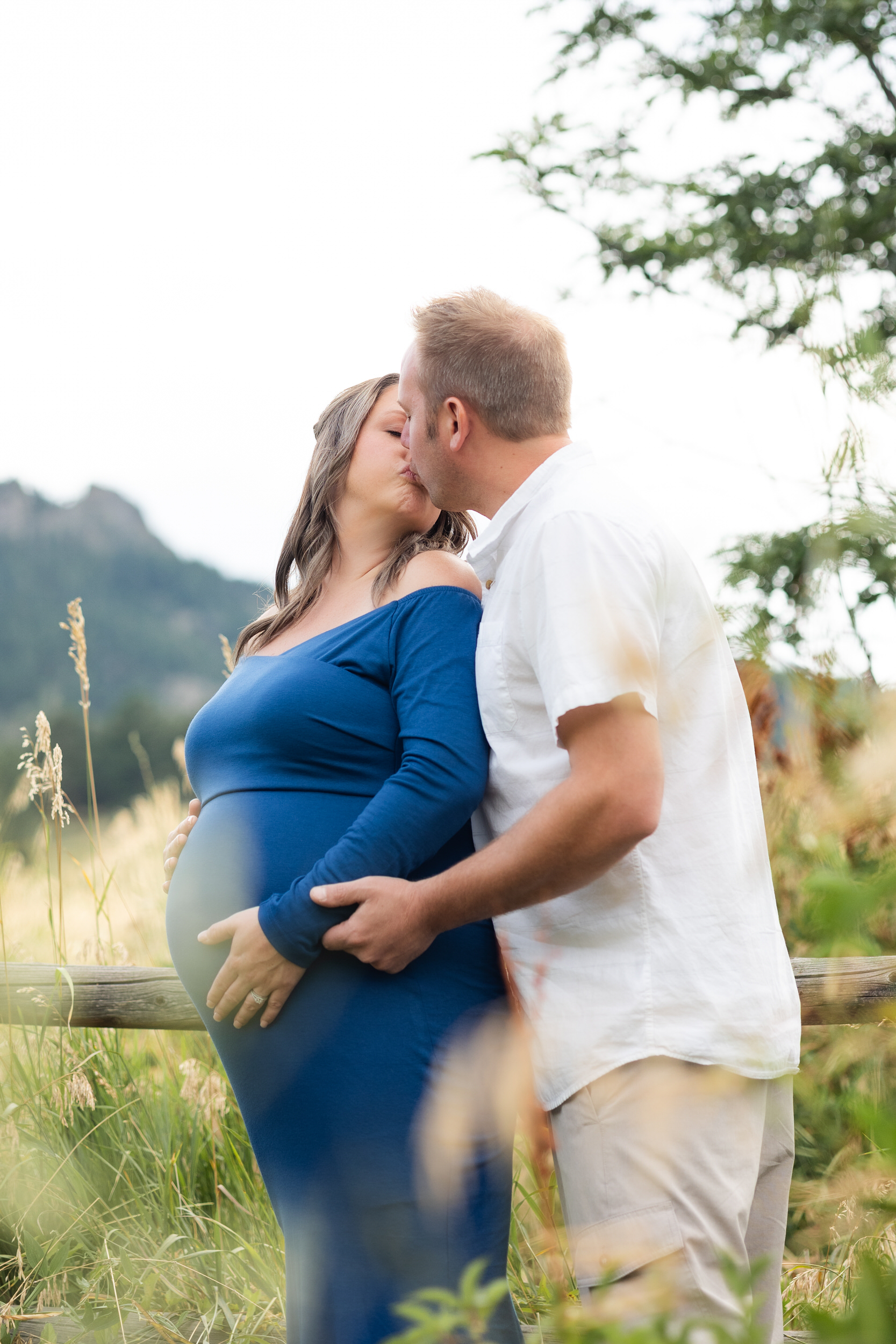 A pregnant woman stands with her back to her husband's chest and they share a kiss while holding her belly.