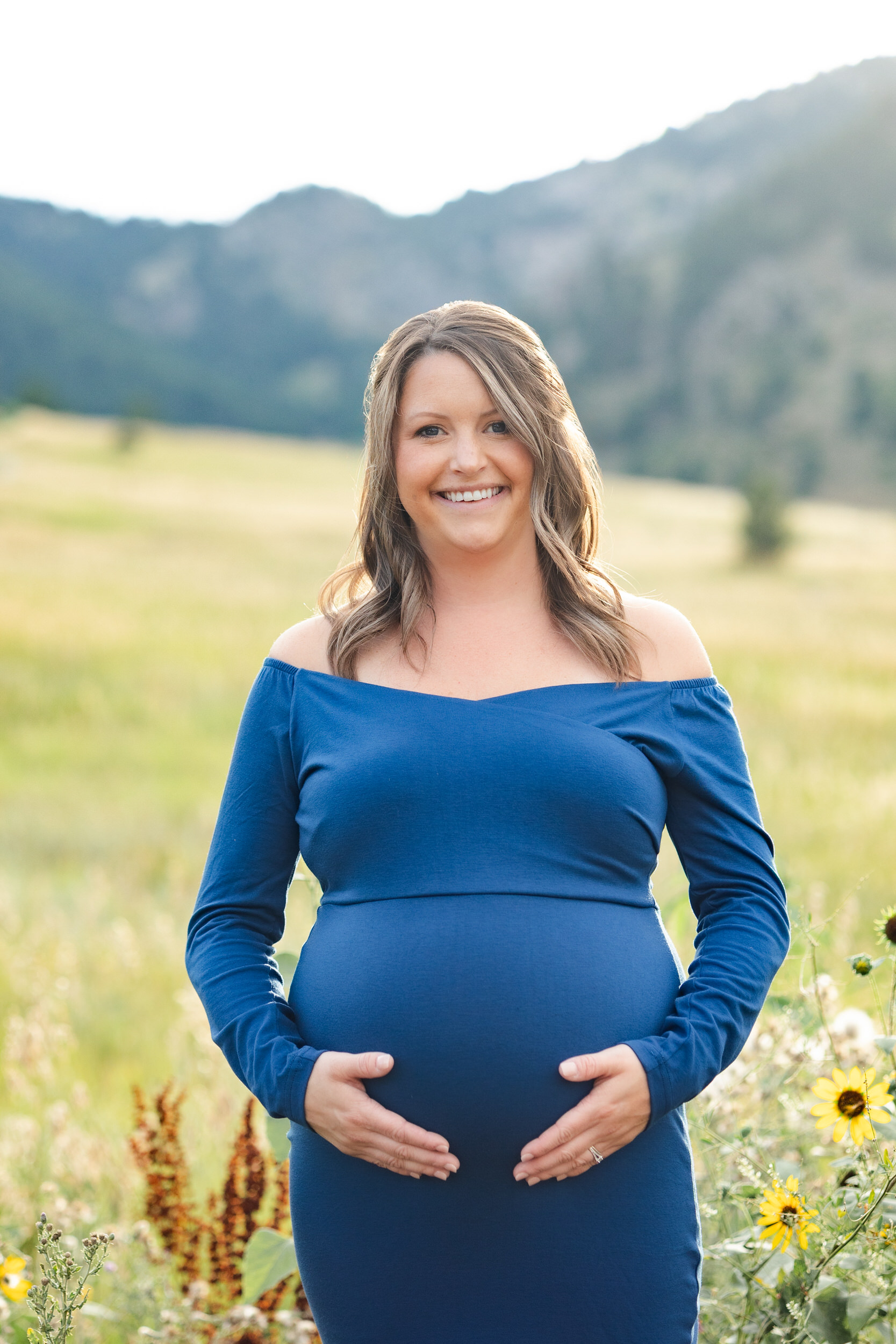 A pregnant woman smiles at the camera with both hands on her belly.
