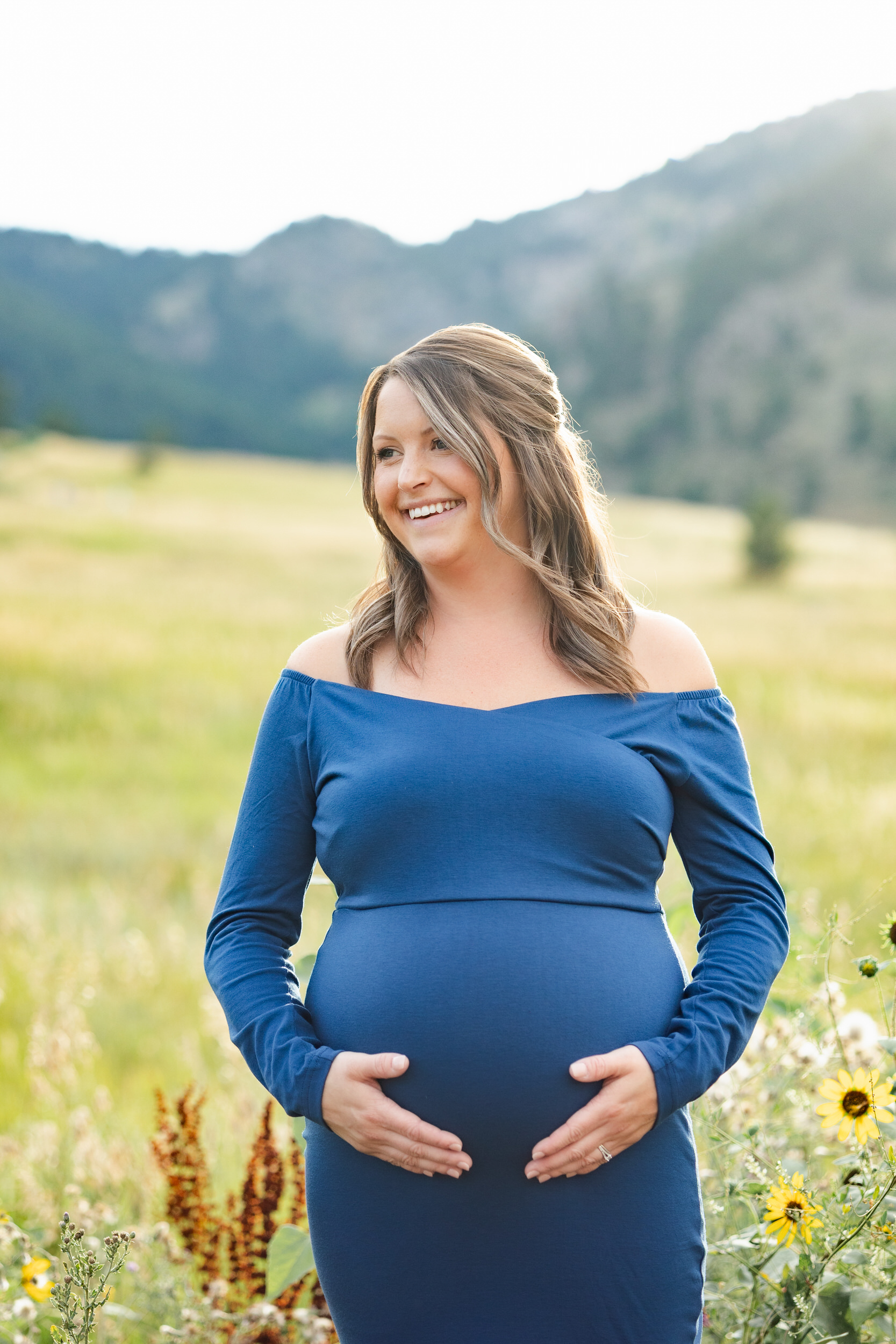 A pregnant woman smiles off camera with both hands on her belly.