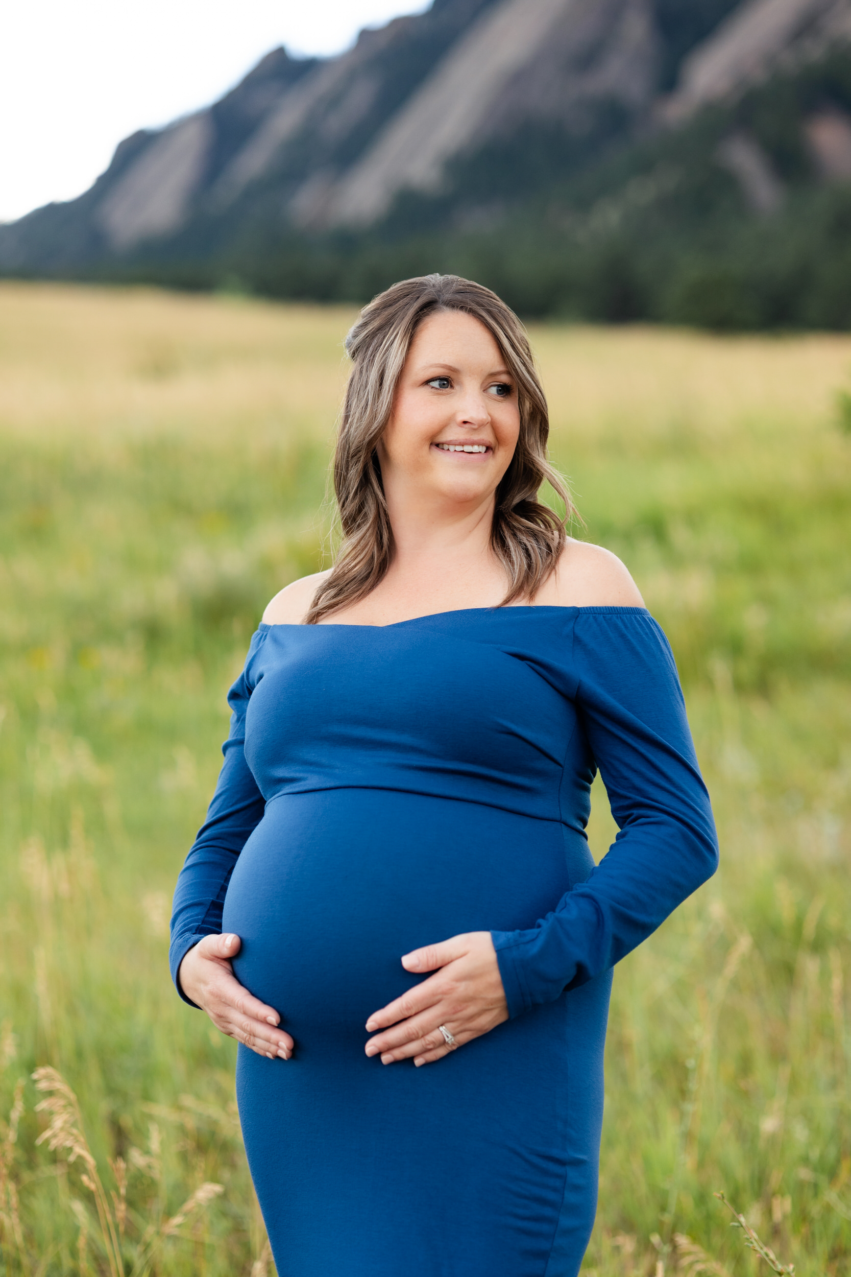 A pregnant woman in a blue dress stands with her hands on her belly and smiles off camera.
