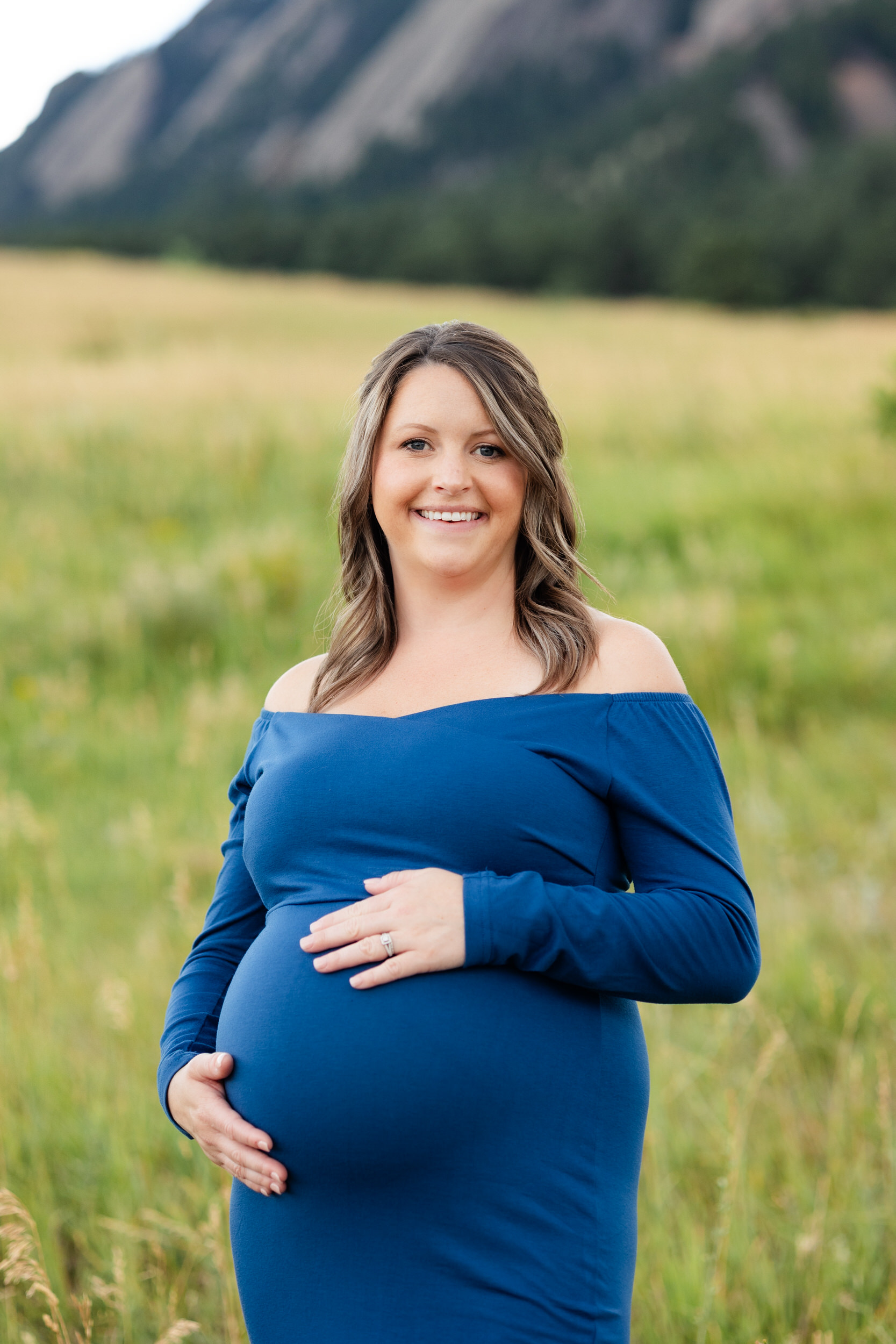 A pregnant woman in a blue dress stands in a field with her hands on her belly and smiles at the camera.