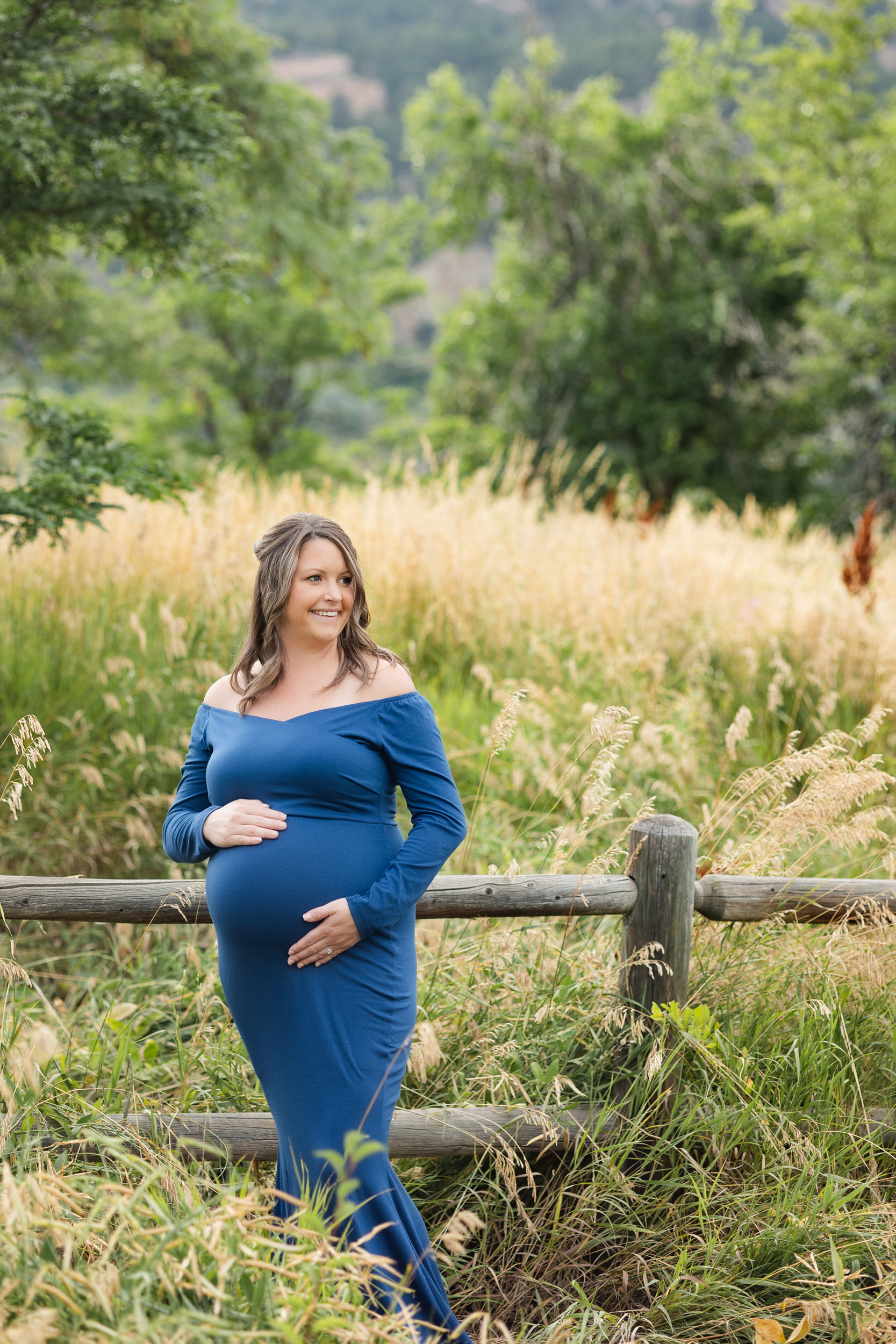 A pregnant woman in a blue dress stands in a field with both hands on her belly and smiles over her shoulder.