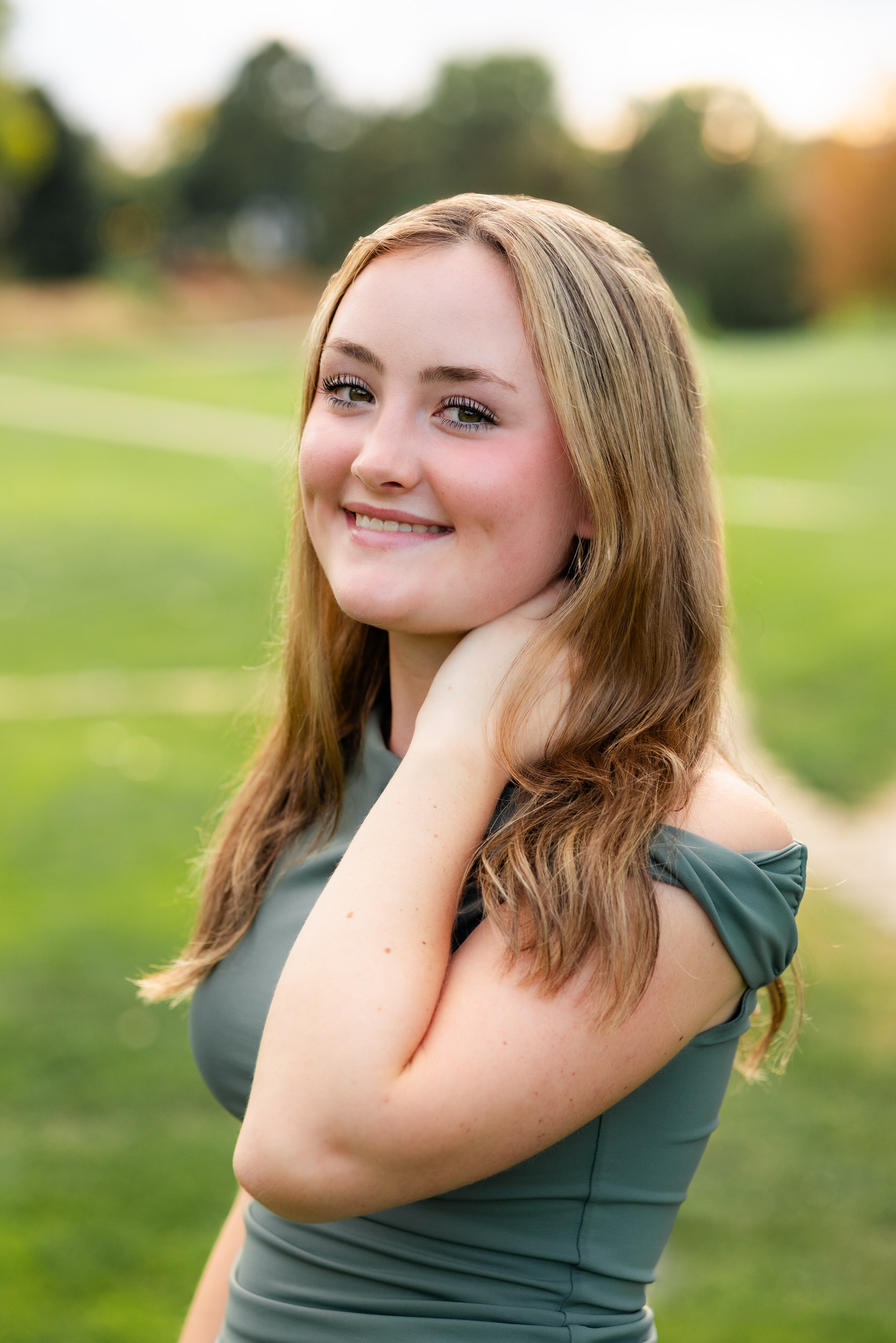 A young woman rests her hand on her neck under her hair and smiles at the camera.