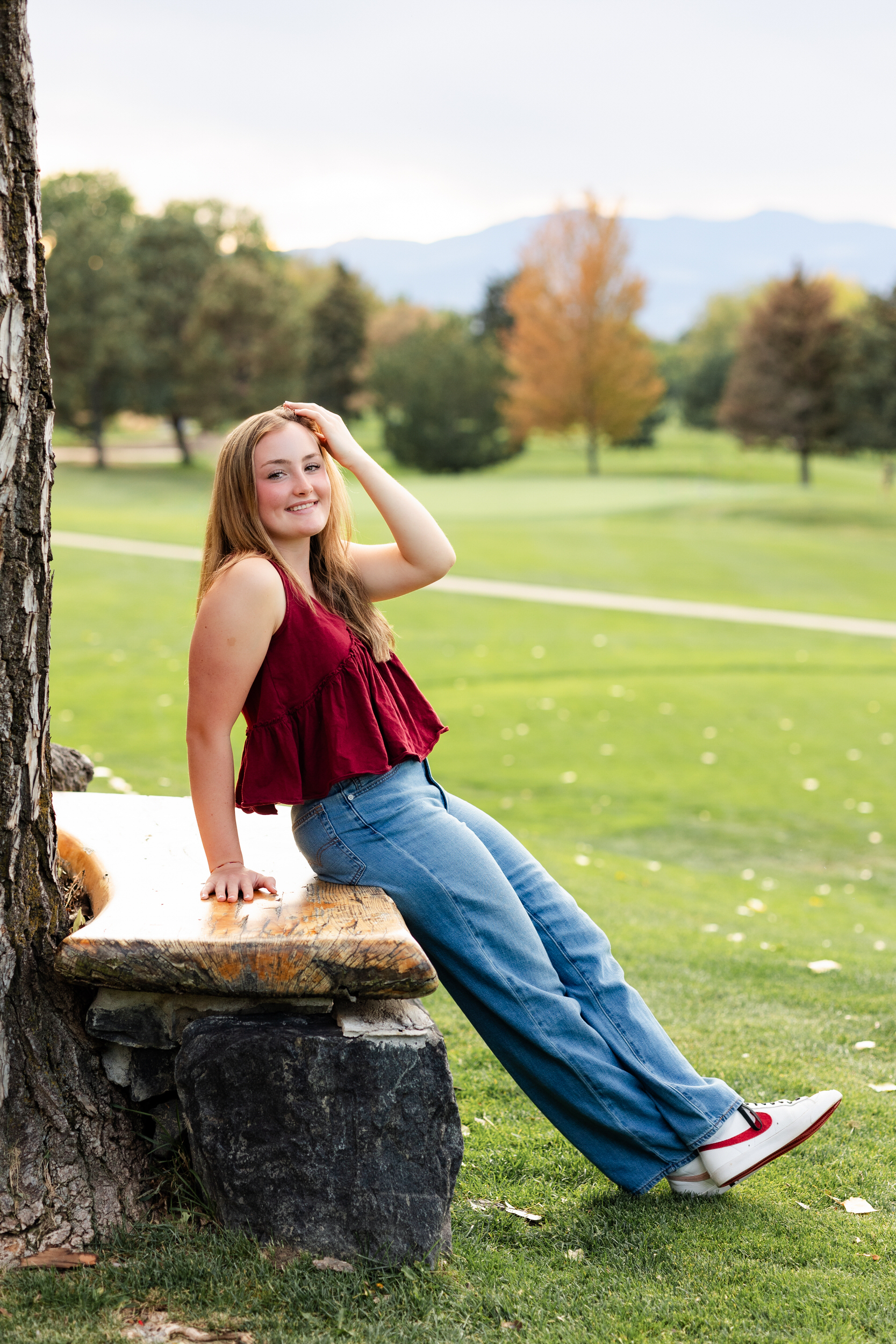 A young woman sitting on a bench runs her hand through her hair and smiles at the camera.