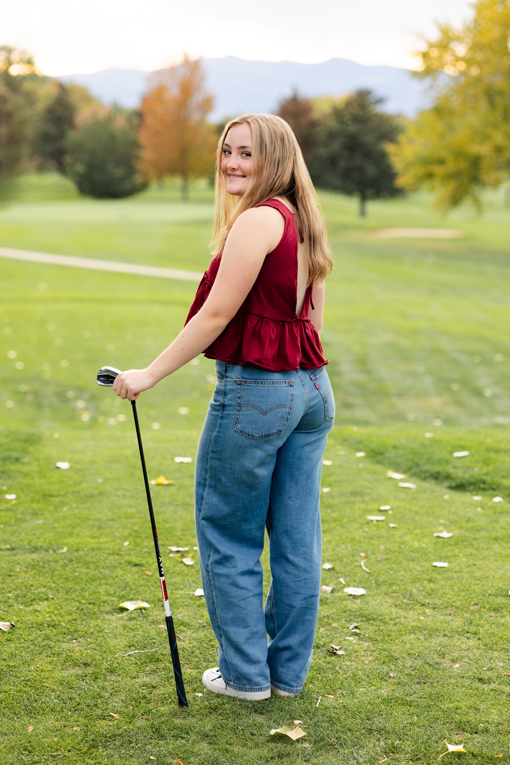 A young woman on a golf course leans on her putter and looks back over her shoulder smiling at the camera.