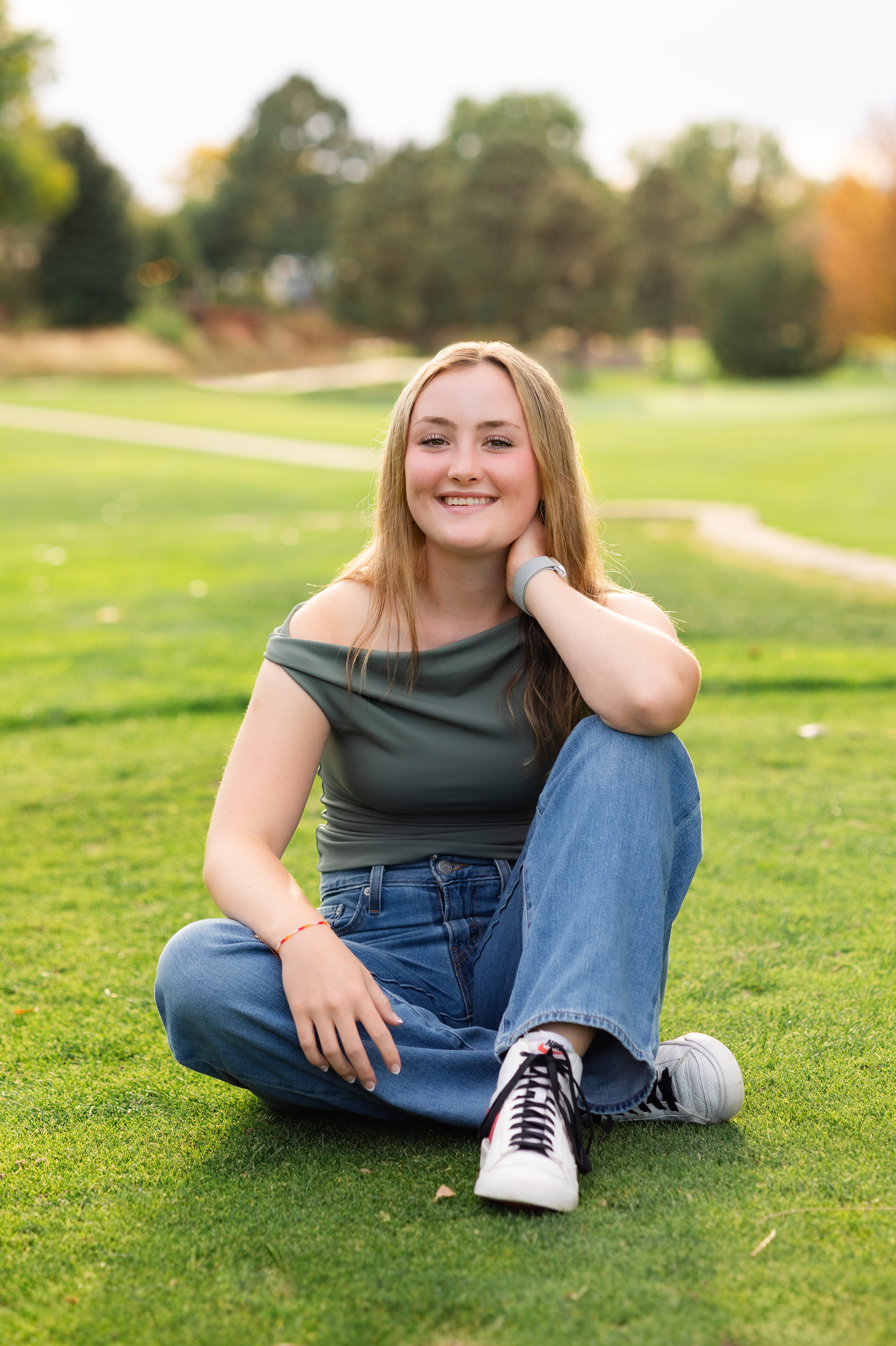 A young woman sits on a golf course resting her elbows on her knees and smiles at the camera.