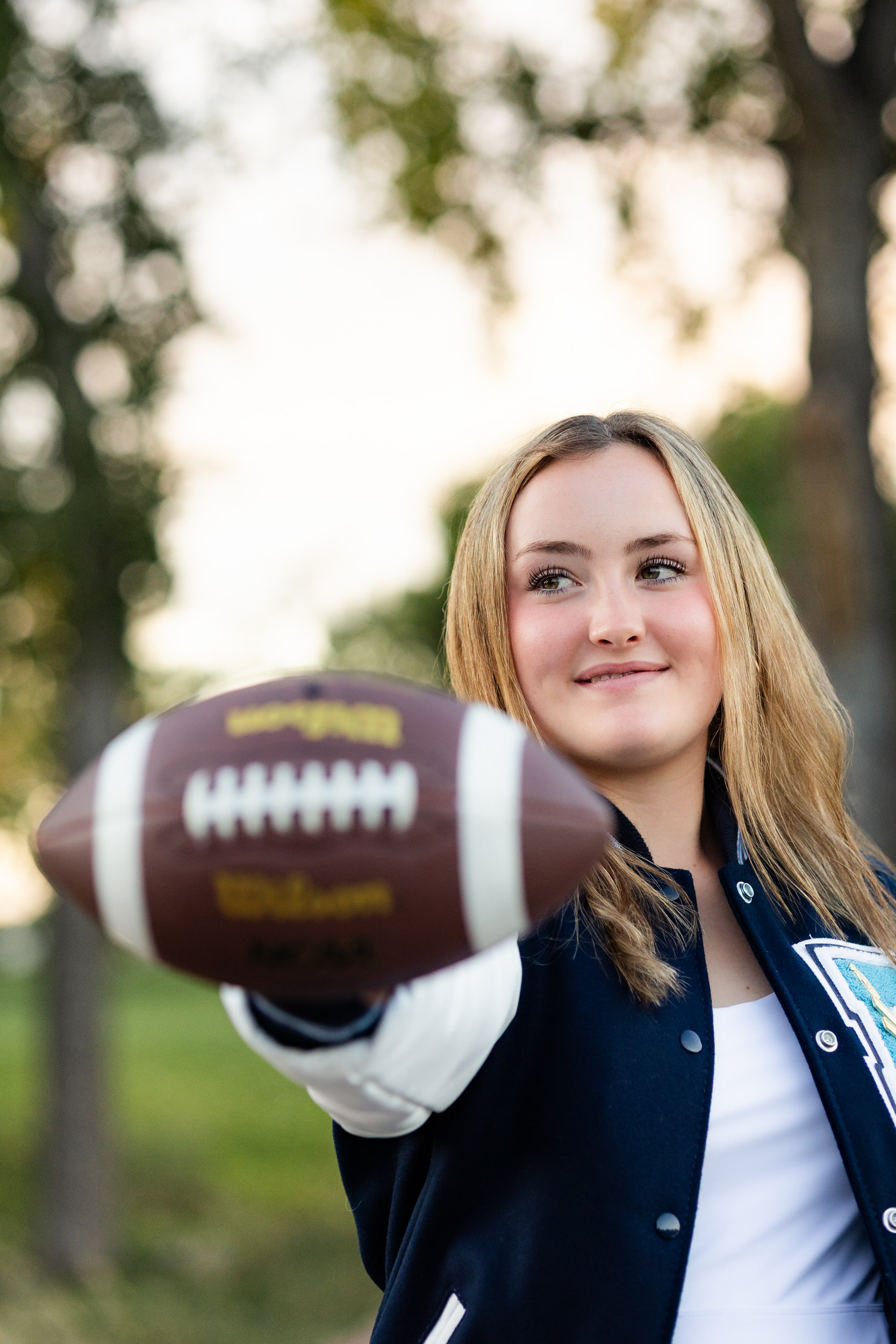 A young woman in a letter jacket holds a football.