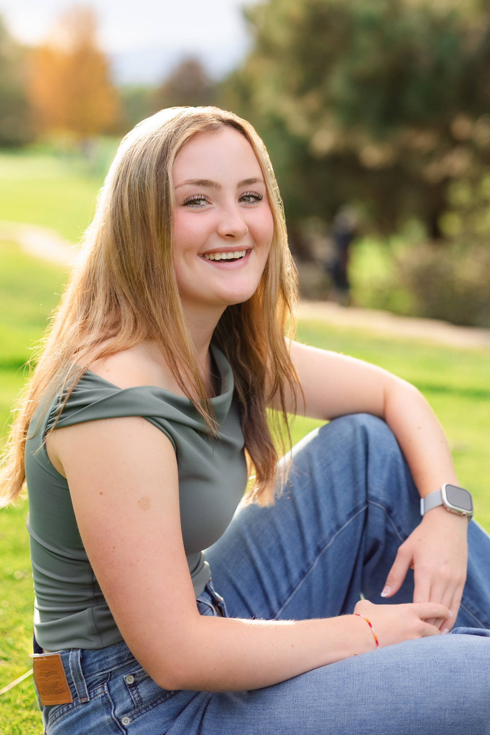 A young woman sits on a golf course resting her elbows on her knees and smiles at the camera.