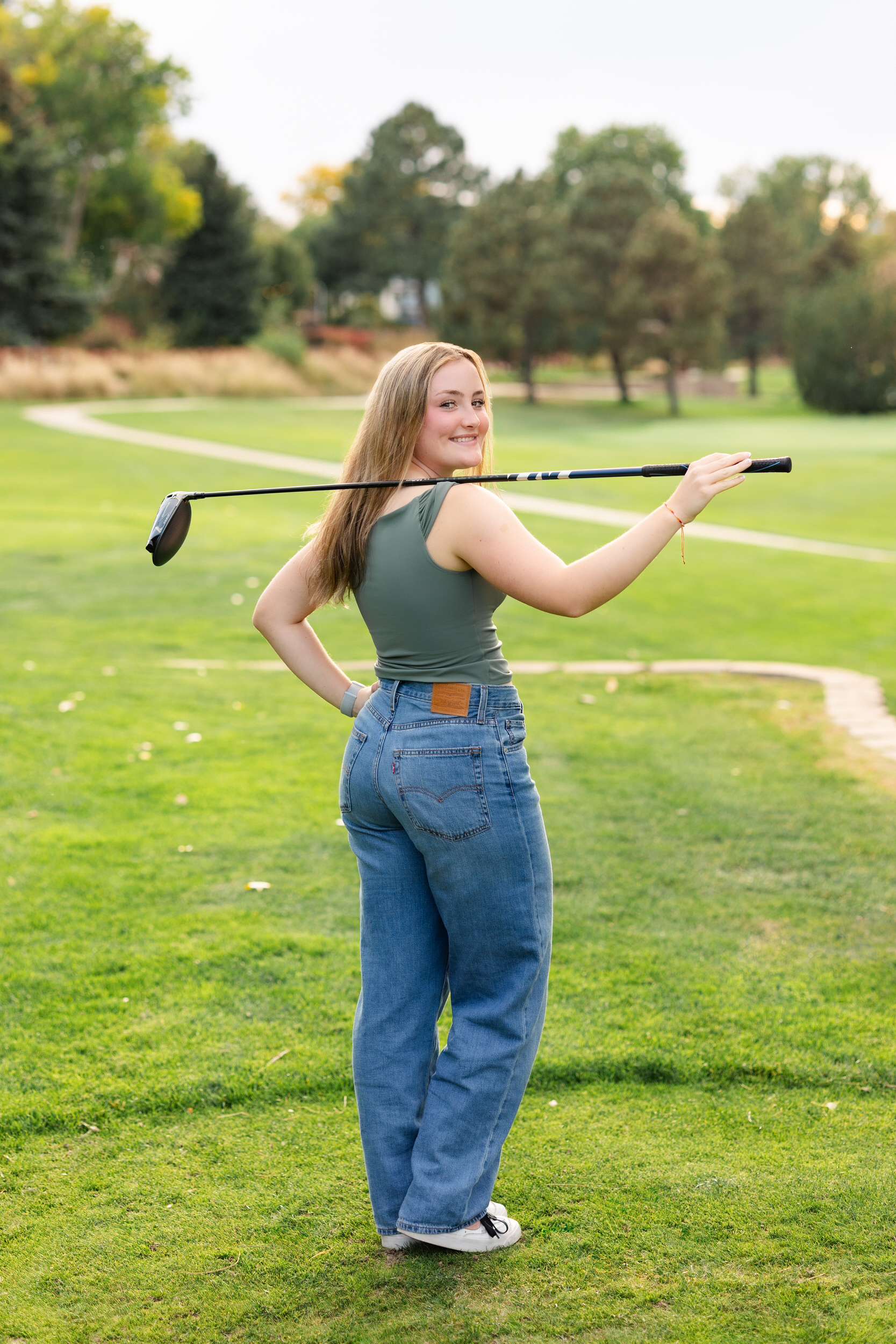 A young woman on a golf course rests a driver on her shoulder and looks back at the camera smiling.