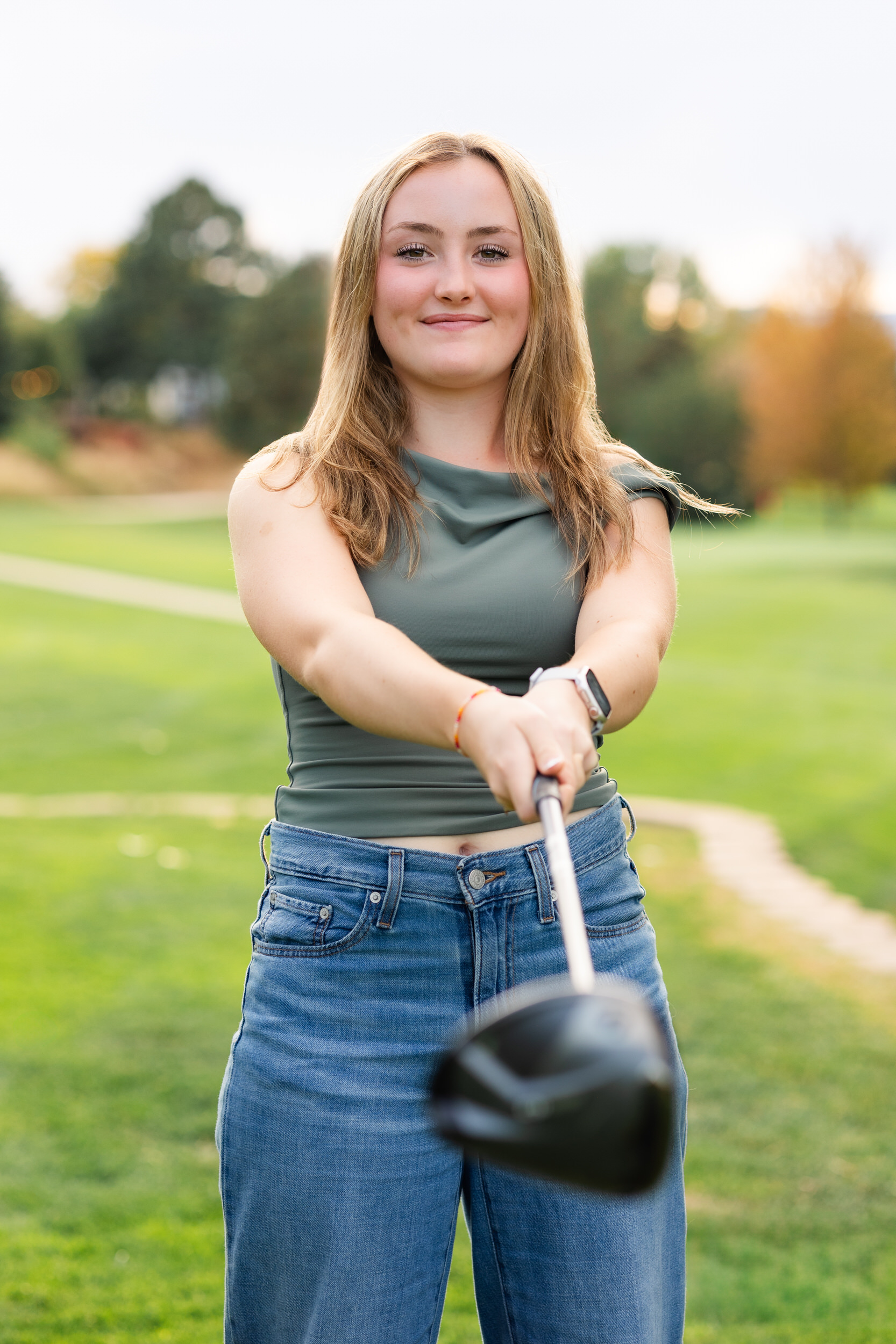 A young woman on a golf course holds a driver toward the camera and smiles.