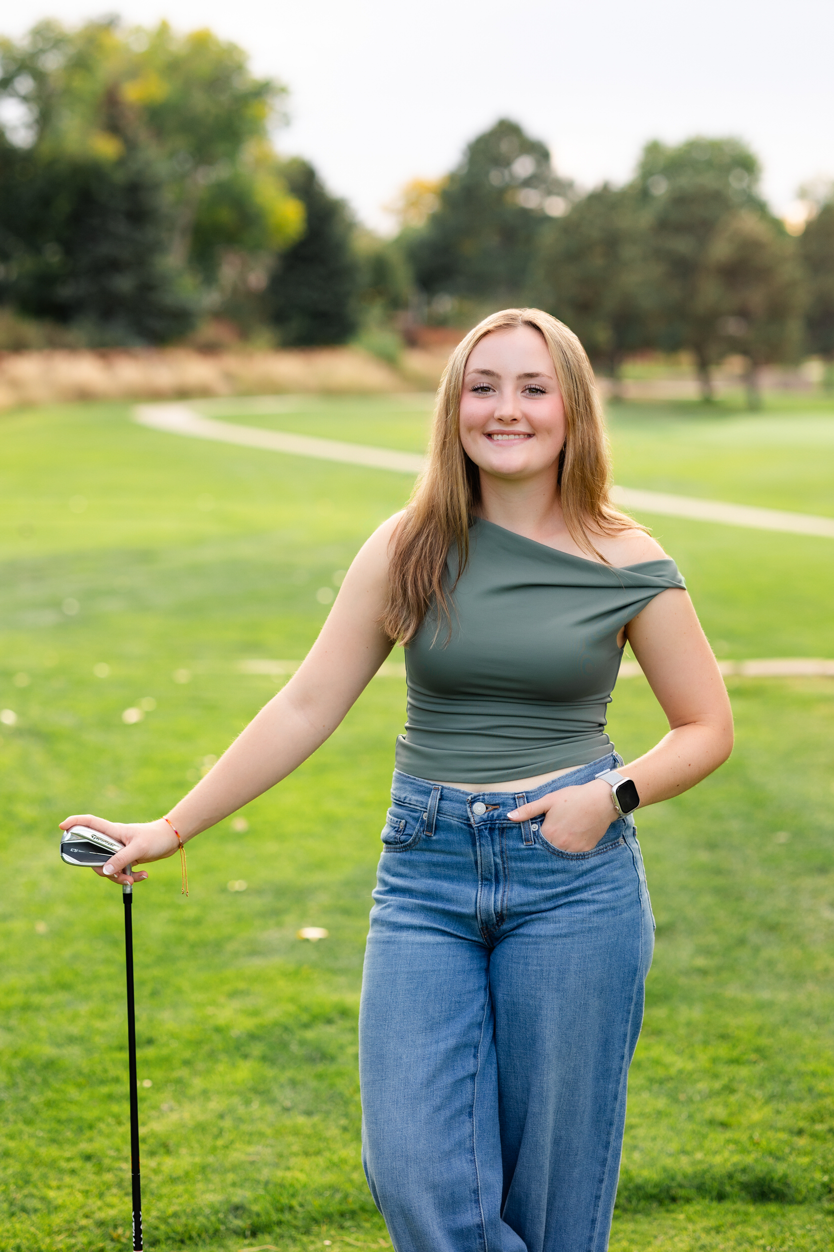 A young woman on a golf course leans on her putter and smiles at the camera.