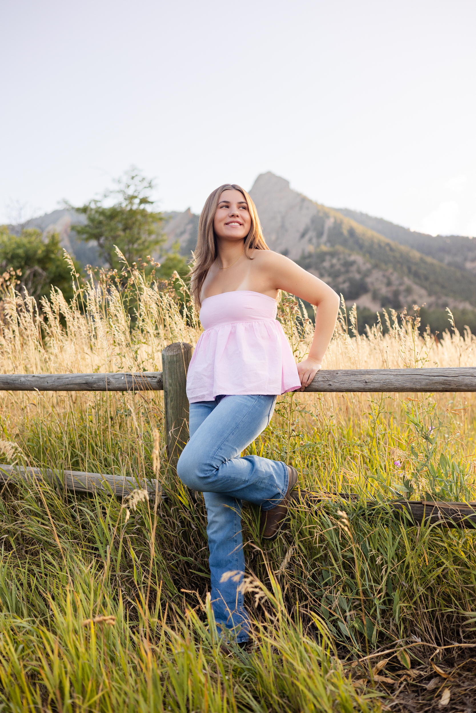 A young woman leans against a wooden fence in a field with mountains behind her.