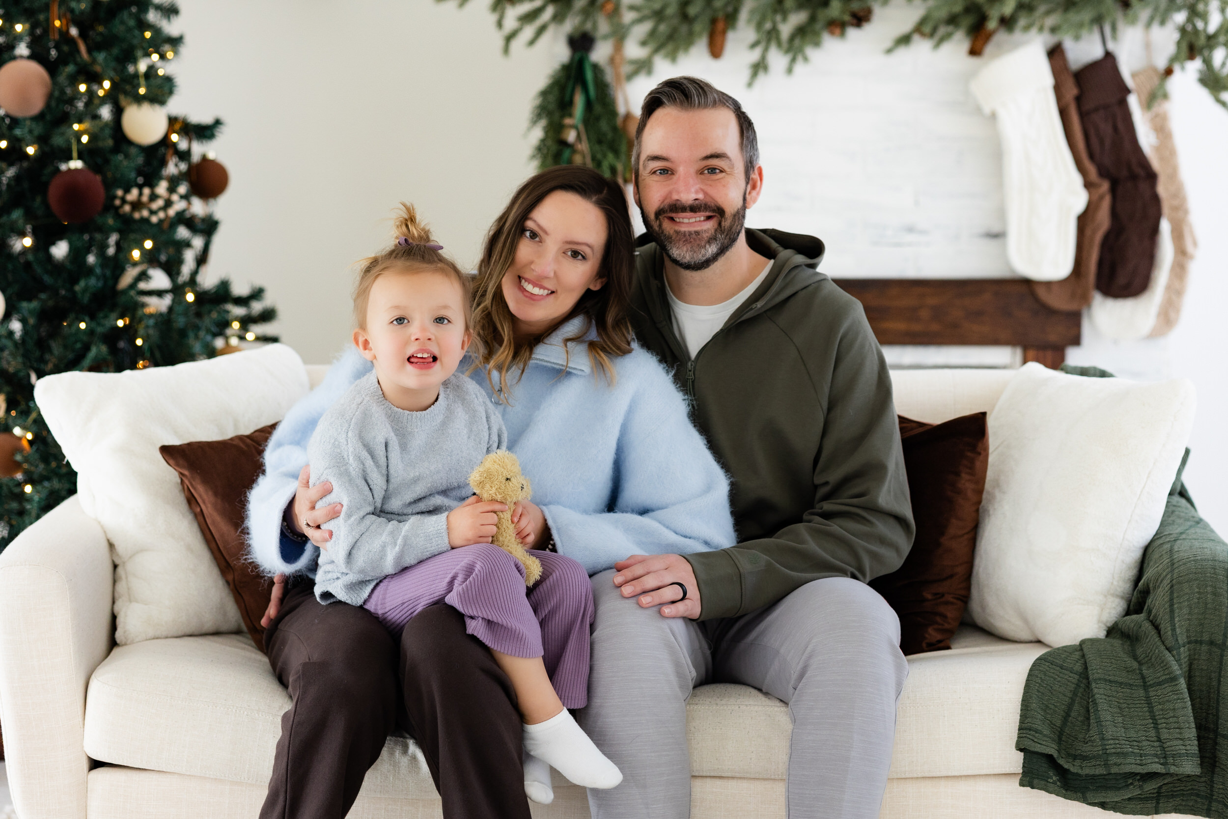 Mom, dad, and young girl sit on a white couch and smile at the camera.