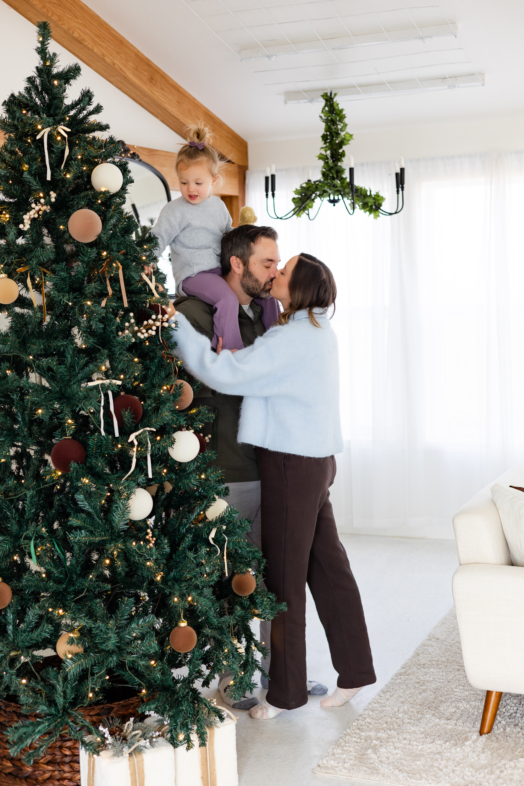 Mom and dad share a kiss next to the Christmas tree while young daughter sits on dad's shoulders and looks at the tree.