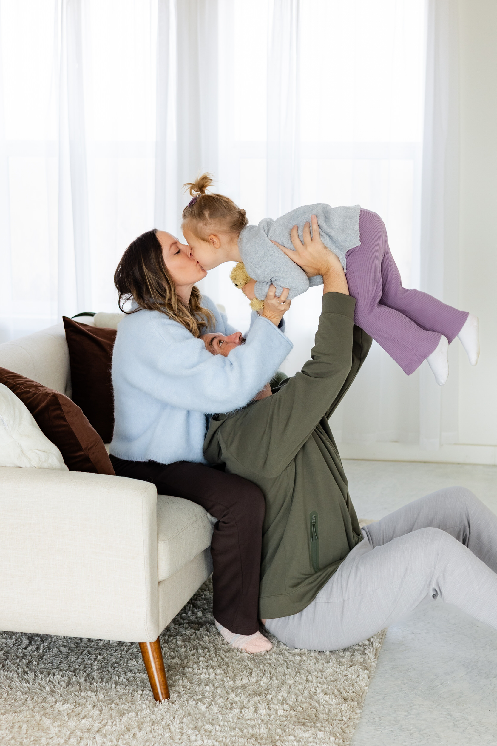 Dad sits on the floor holding young daughter in the air while mom sits on a couch and gives young daughter a kiss.