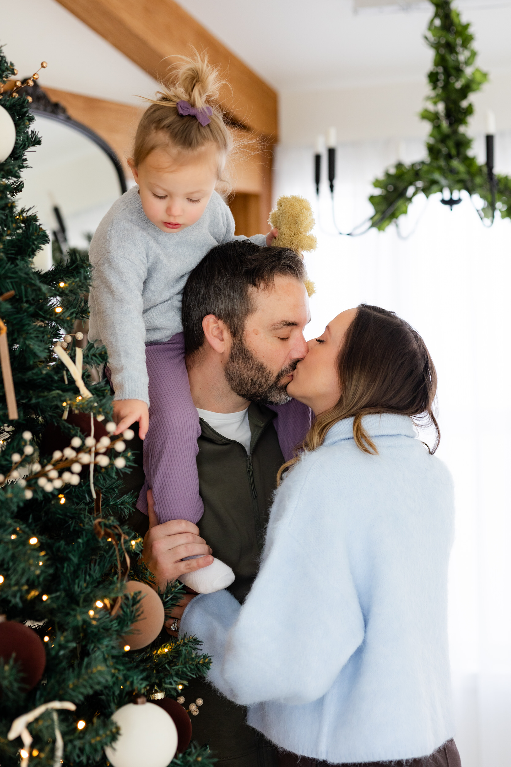 Dad holds his young daughter on his shoulders while he kisses his wife.