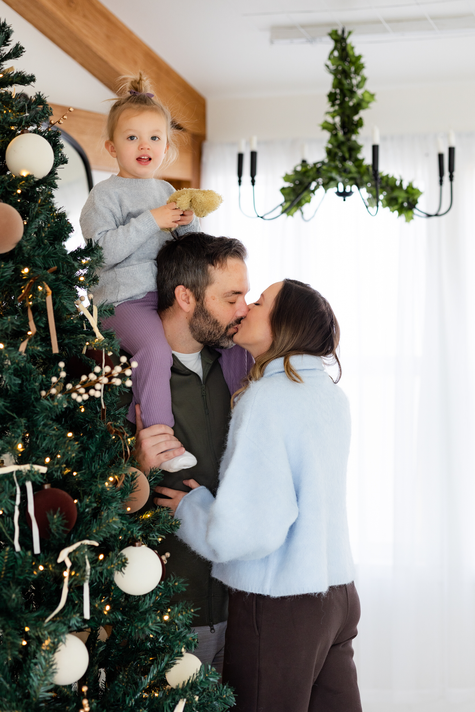 Little girl sits on her dad's shoulders and smiles at camera while mom and dad share a kiss.