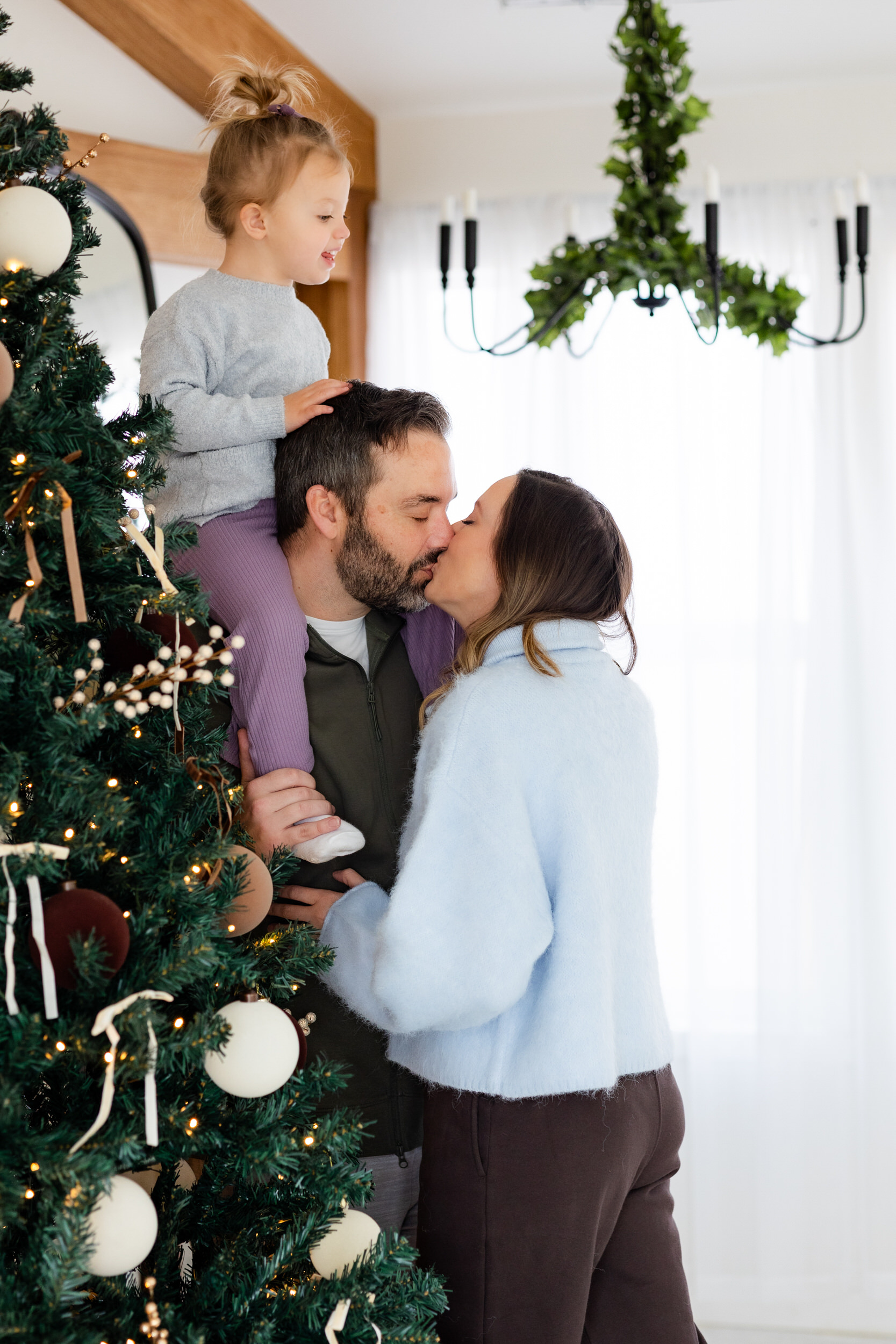Little girl sits on her dad's shoulders and smiles down at her parents who are sharing a kiss by the Christmas tree.
