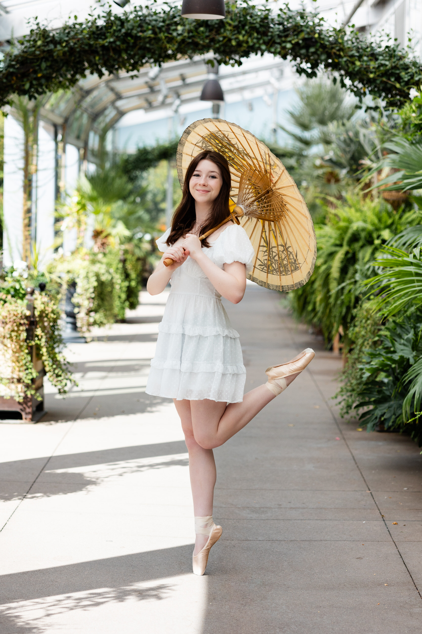 A young woman holding a parasol on one shoulder stands en pointe in a greenhouse at the Denver Botanic Gardens.