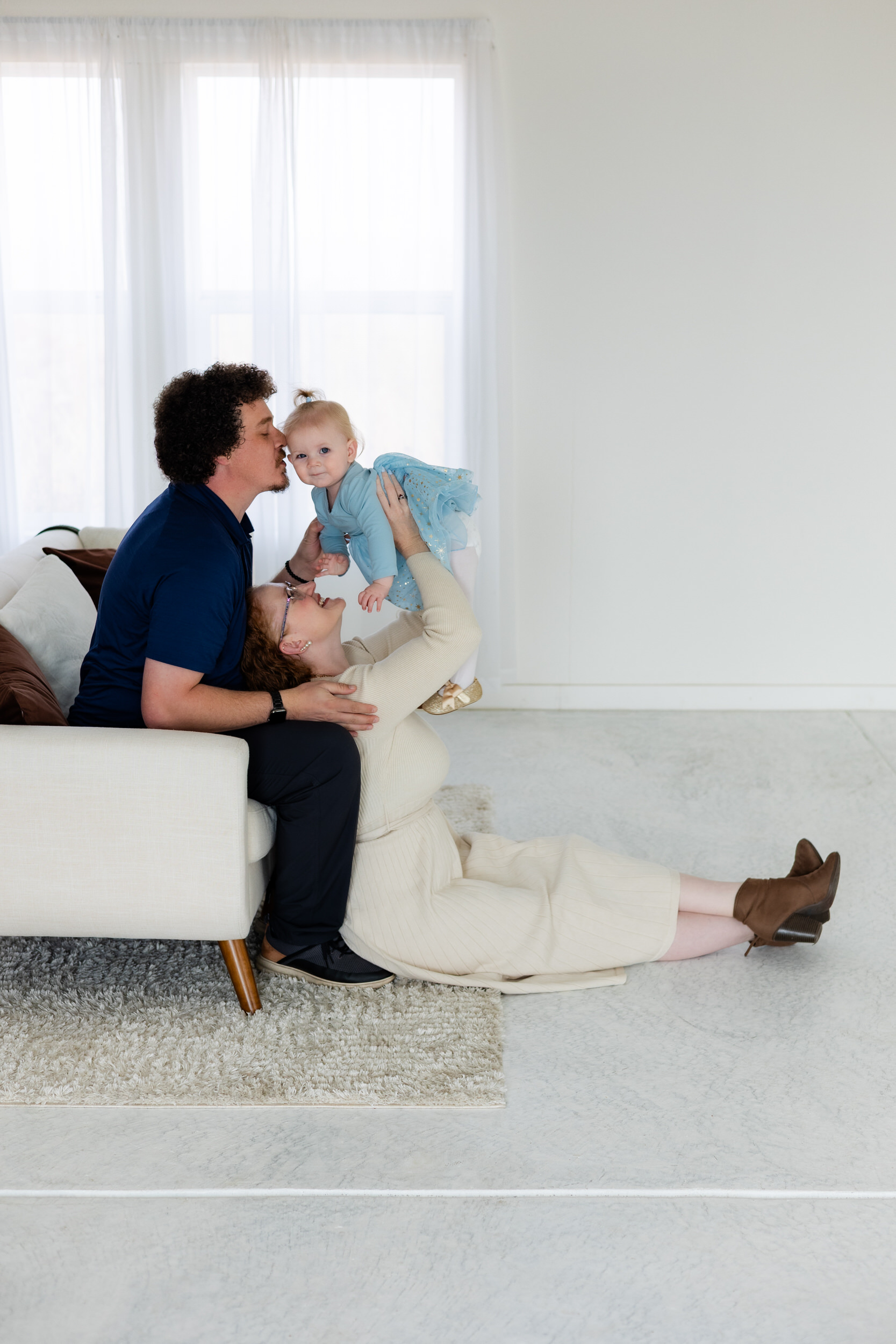 Dad is sitting on couch while mom sits on floor with her back to his legs while she holds baby girl up in the air as dad kisses her cheek.