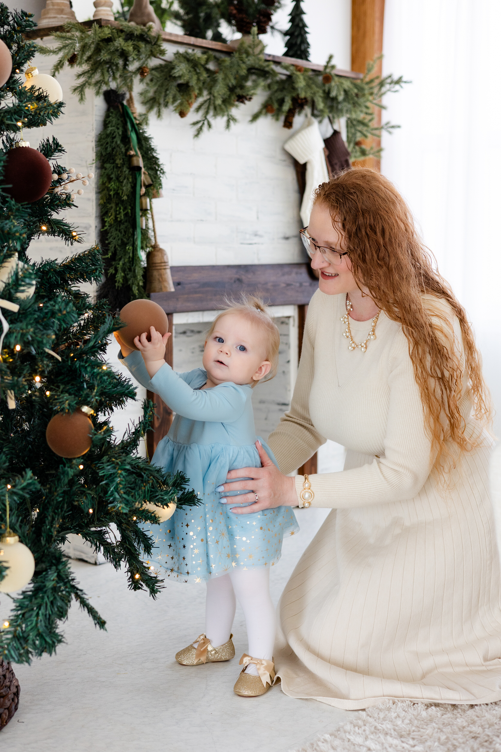 Baby girl touches ornament on Christmas tree while mom holds her.