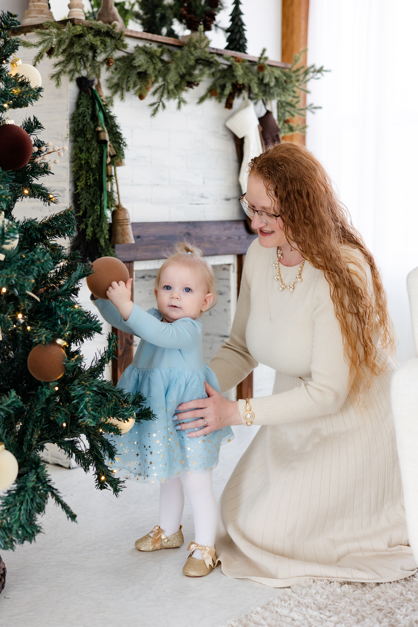 Baby girl touches ornament on Christmas tree while mom holds her.