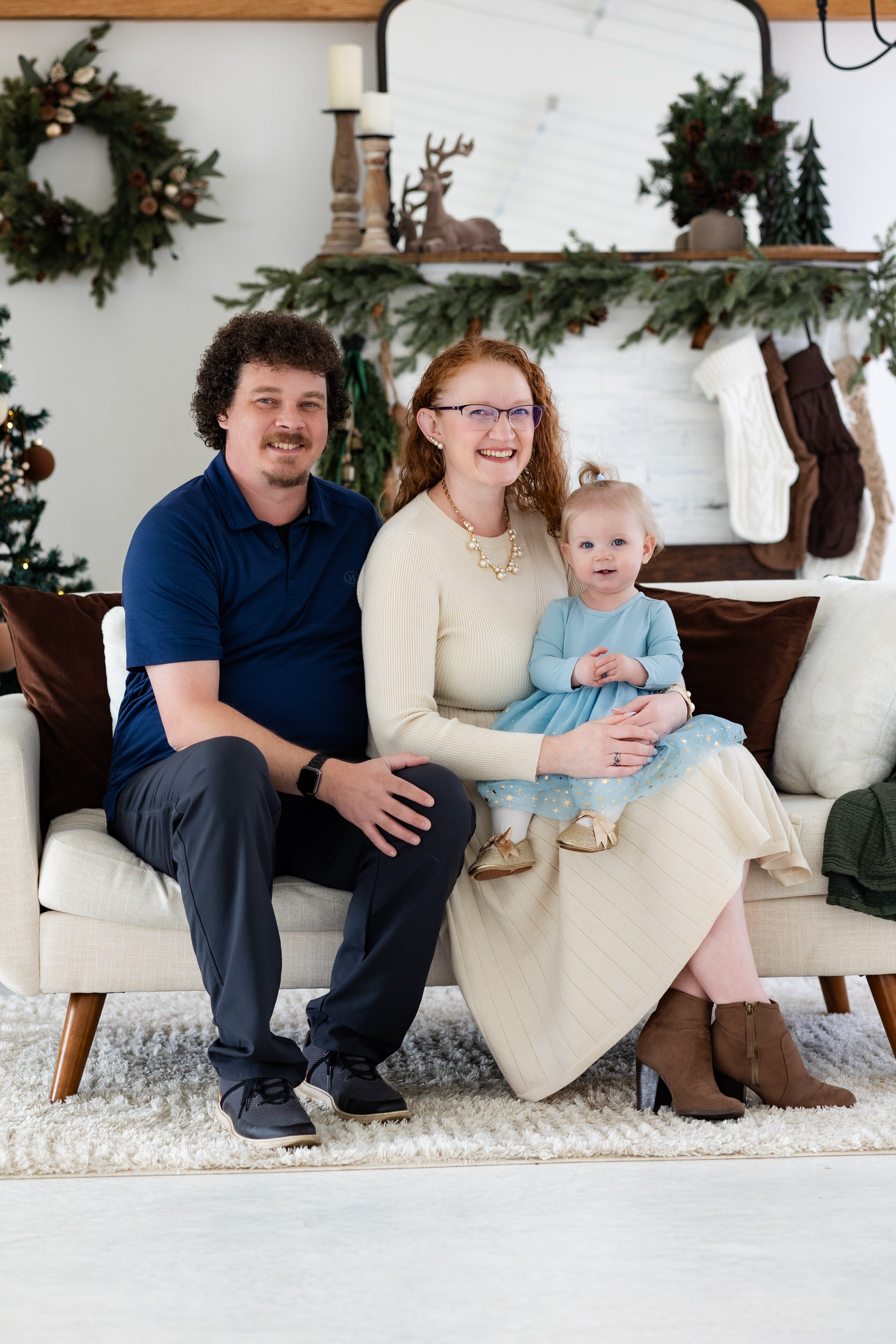 Mom and dad sit on a white couch holding their baby girl and smiling at the camera.