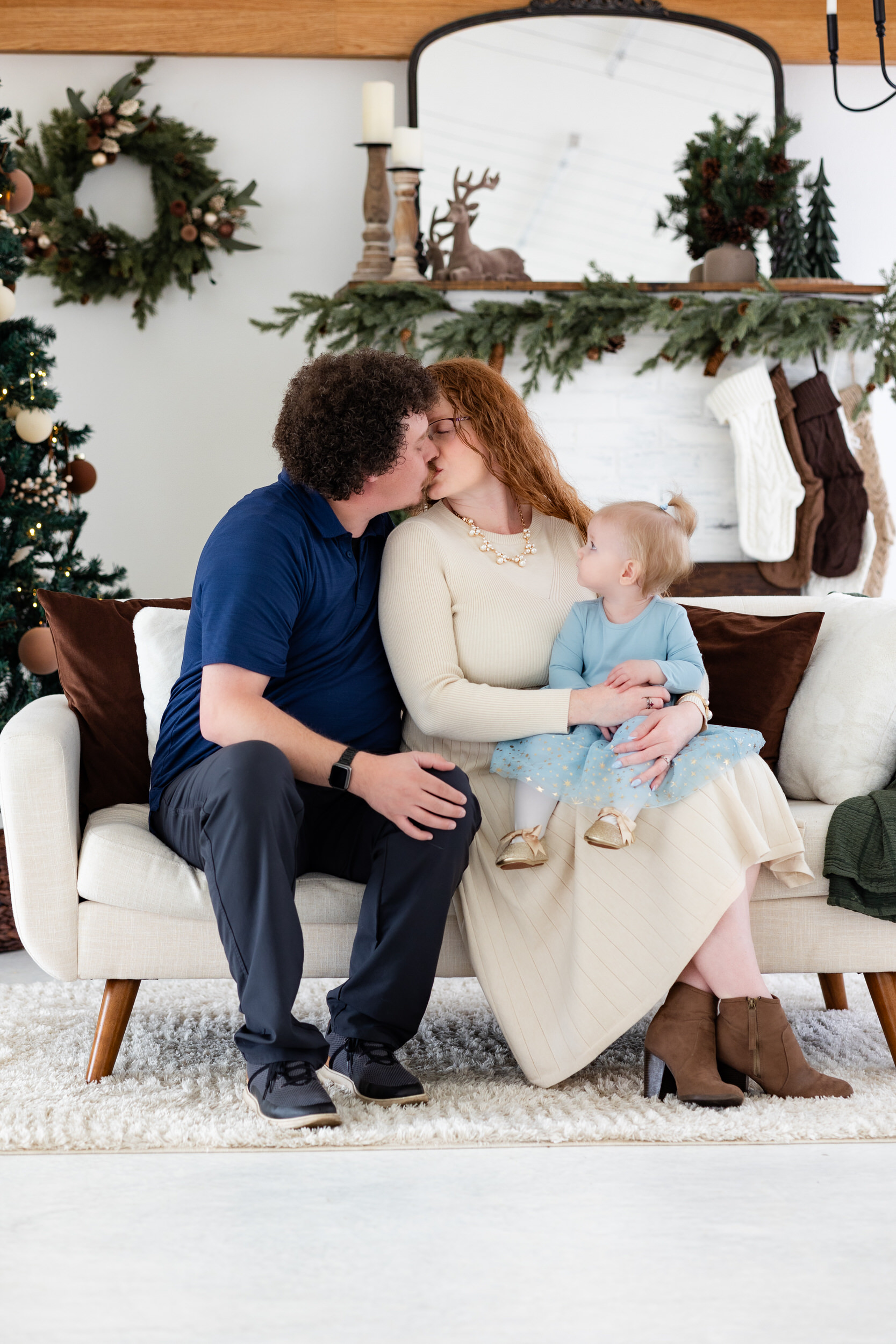 Mom and dad sit on a couch and share a kiss while baby girl looks at them.