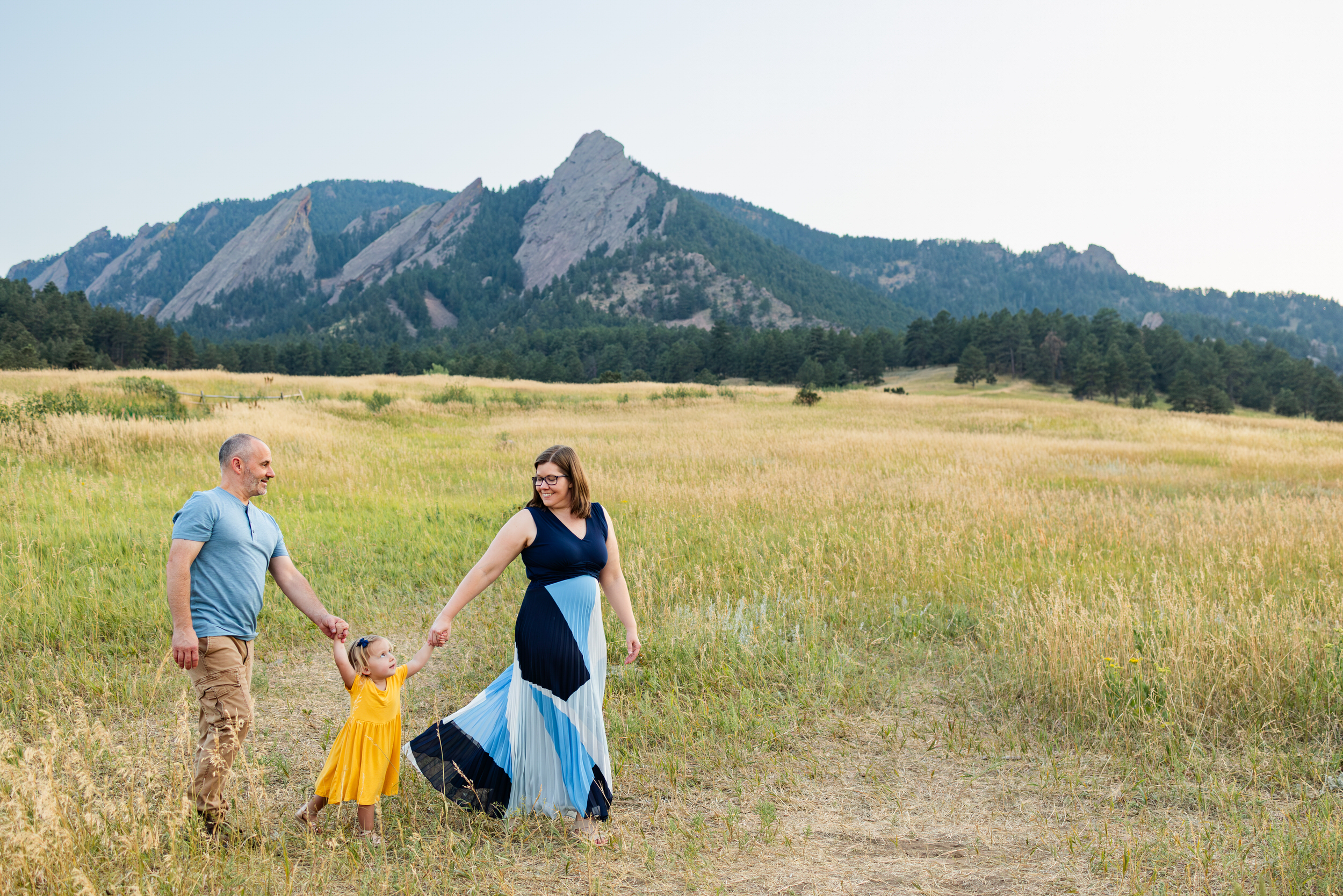 A mom and dad hold hands with their toddler daughter and walk across a field in the mountains.