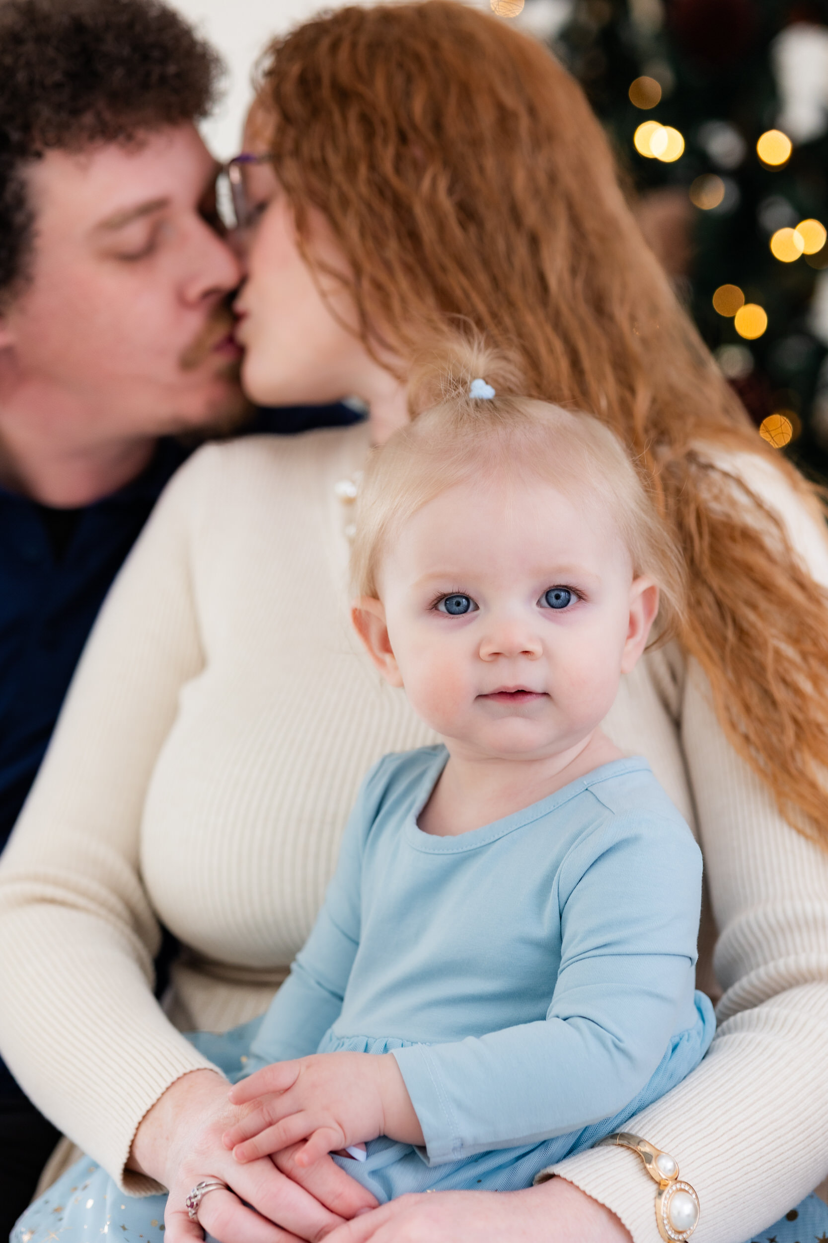 Mom and dad kiss in the background while baby girl looks at the camera.