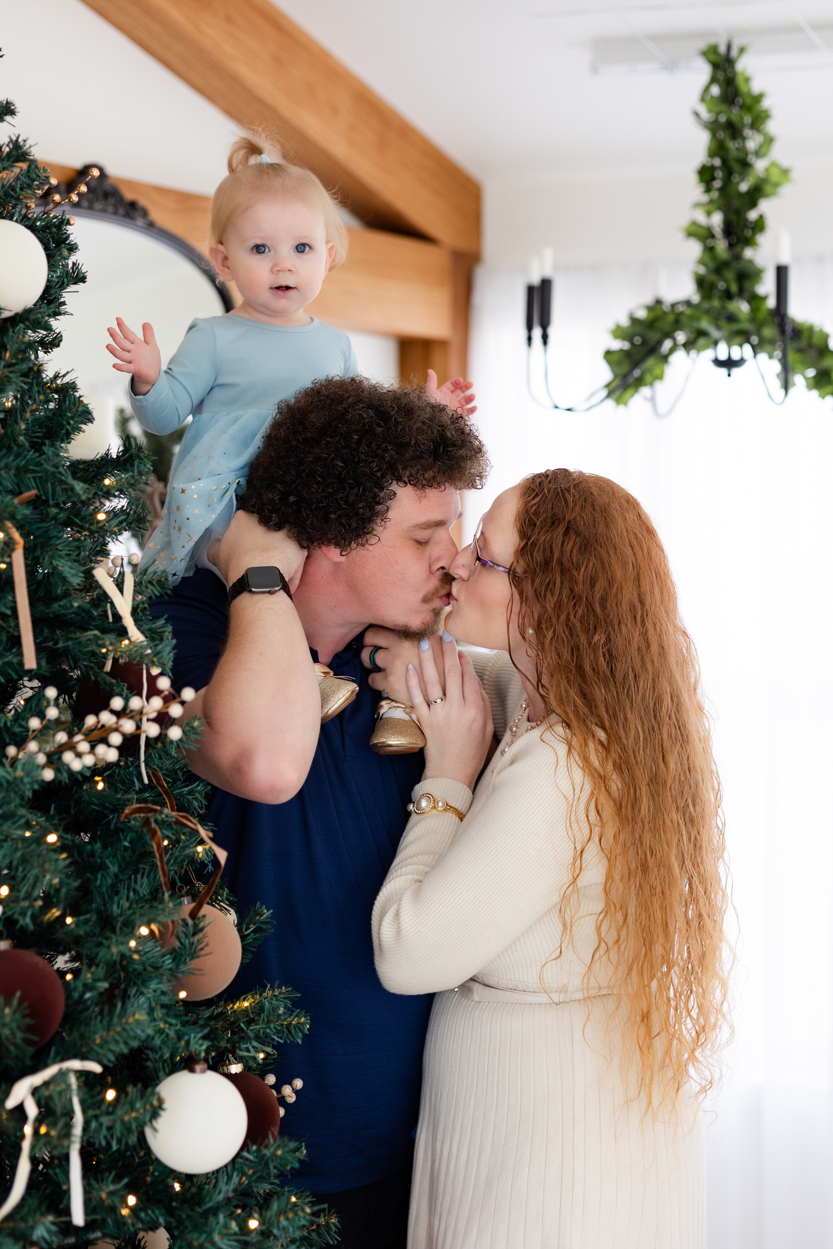 Dad holds baby girl on his shoulders while he and his wife share a kiss.