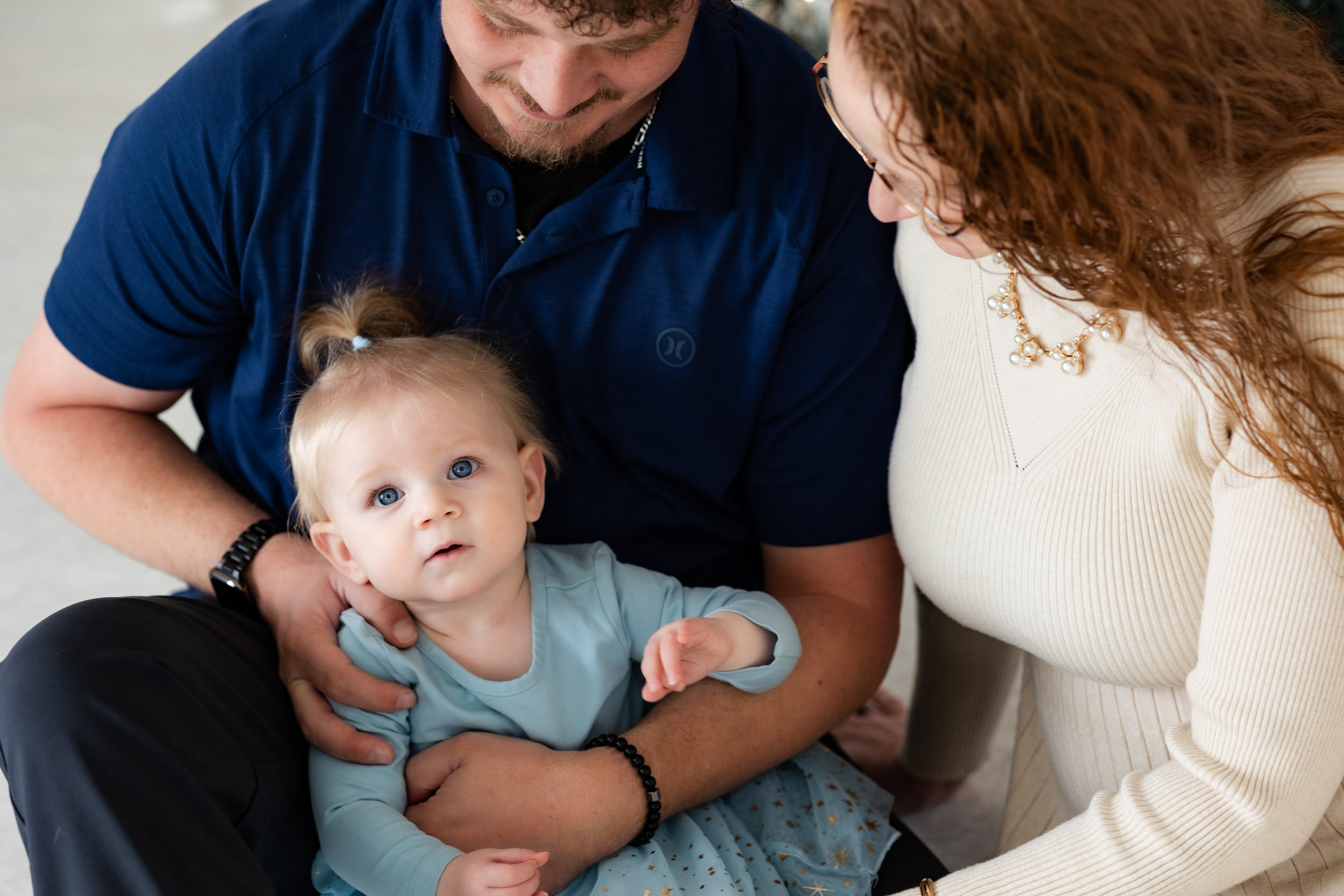 Mom and dad look at baby girl while baby girl looks at camera.