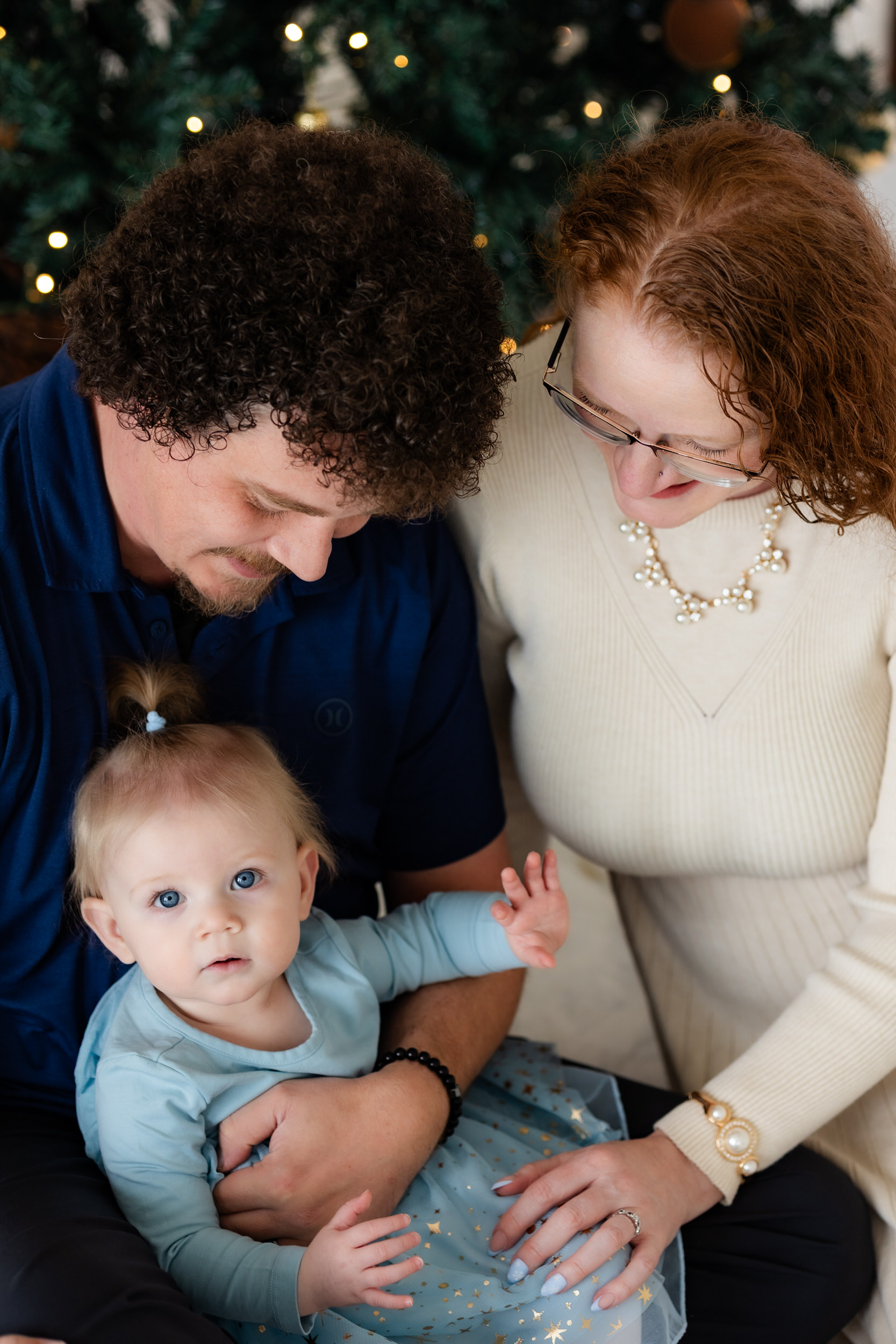 Mom and dad look at baby girl while baby girl looks at camera.