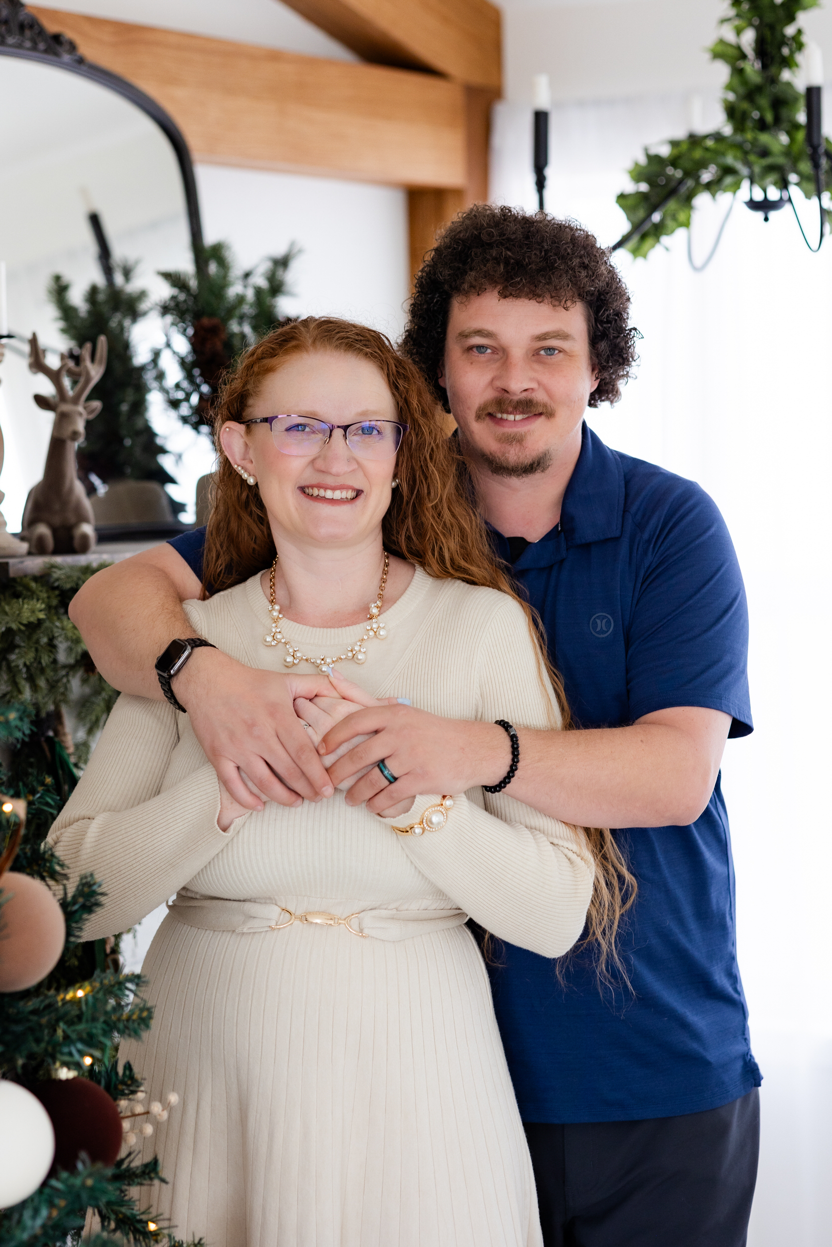 Husband hugs his wife from behind around her shoulders and they both smile at the camera.
