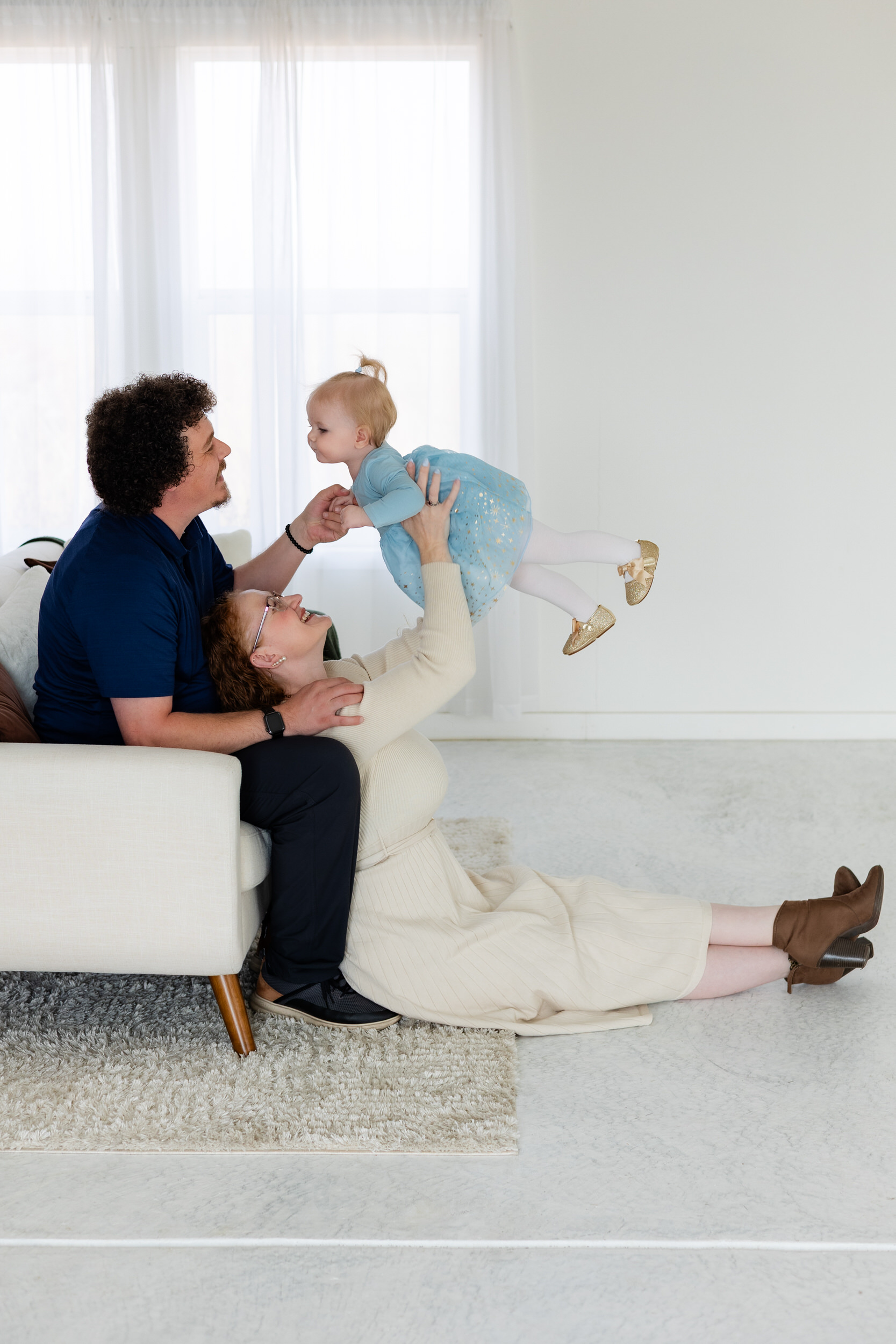 Dad is sitting on couch while mom sits on floor with her back to his legs while she holds baby girl up in the air to look at dad.