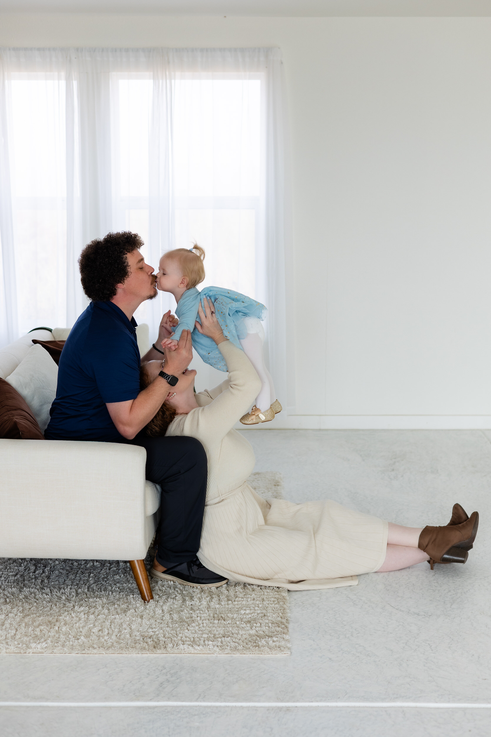 Dad is sitting on couch while mom sits on floor with her back to his legs while she holds baby girl up in the air to kiss dad.