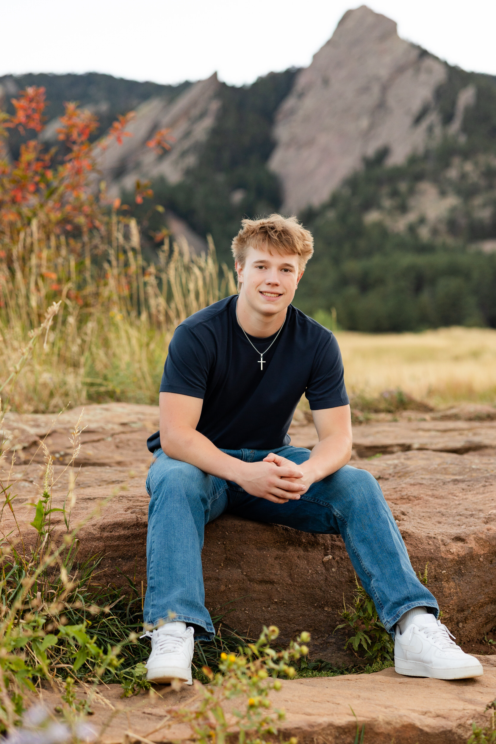A young man sits on a rock and smiles at the camera.