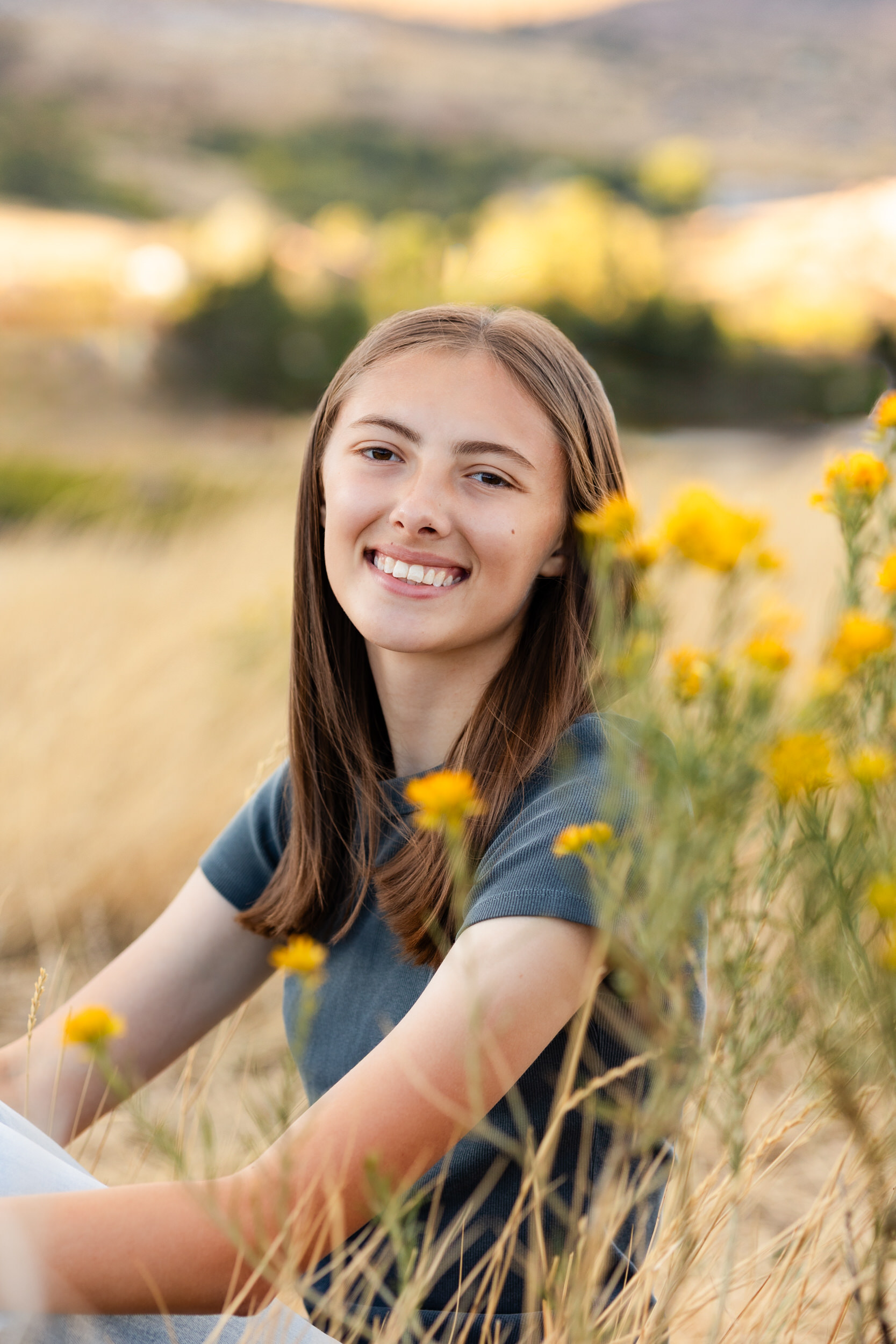 A young woman sits in a field behind yellow flowers and smiles at the camera.