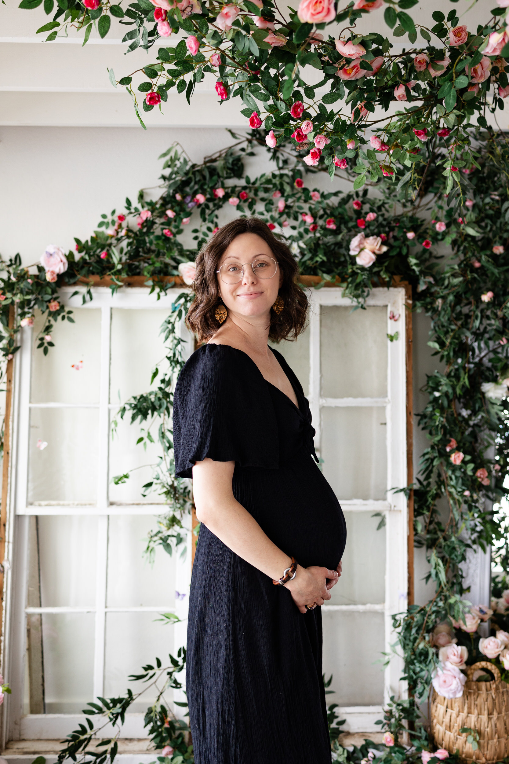 A pregnant woman stands in front of a wall of flowers.