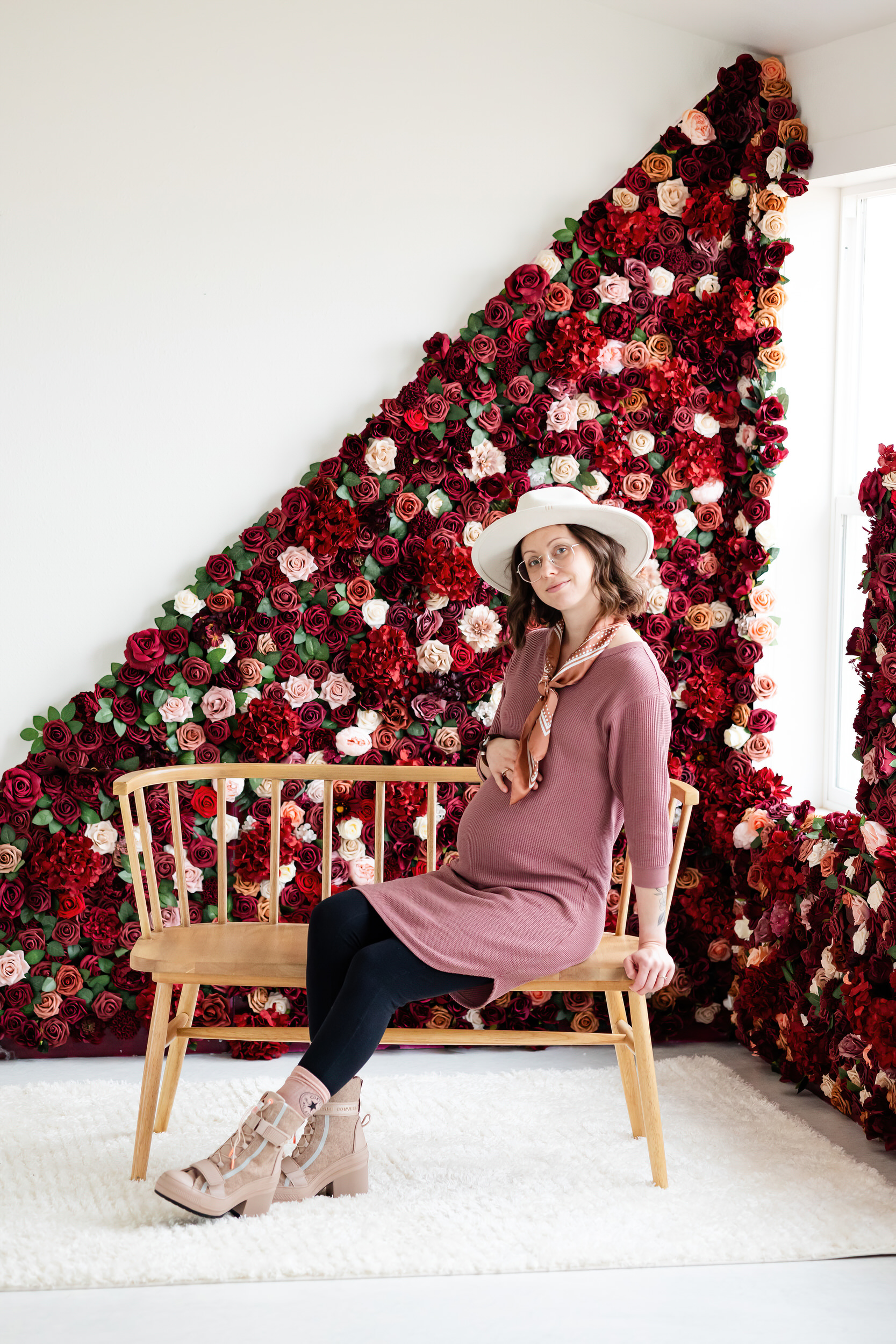 A pregnant woman sits on a bench in front of a wall of flowers.