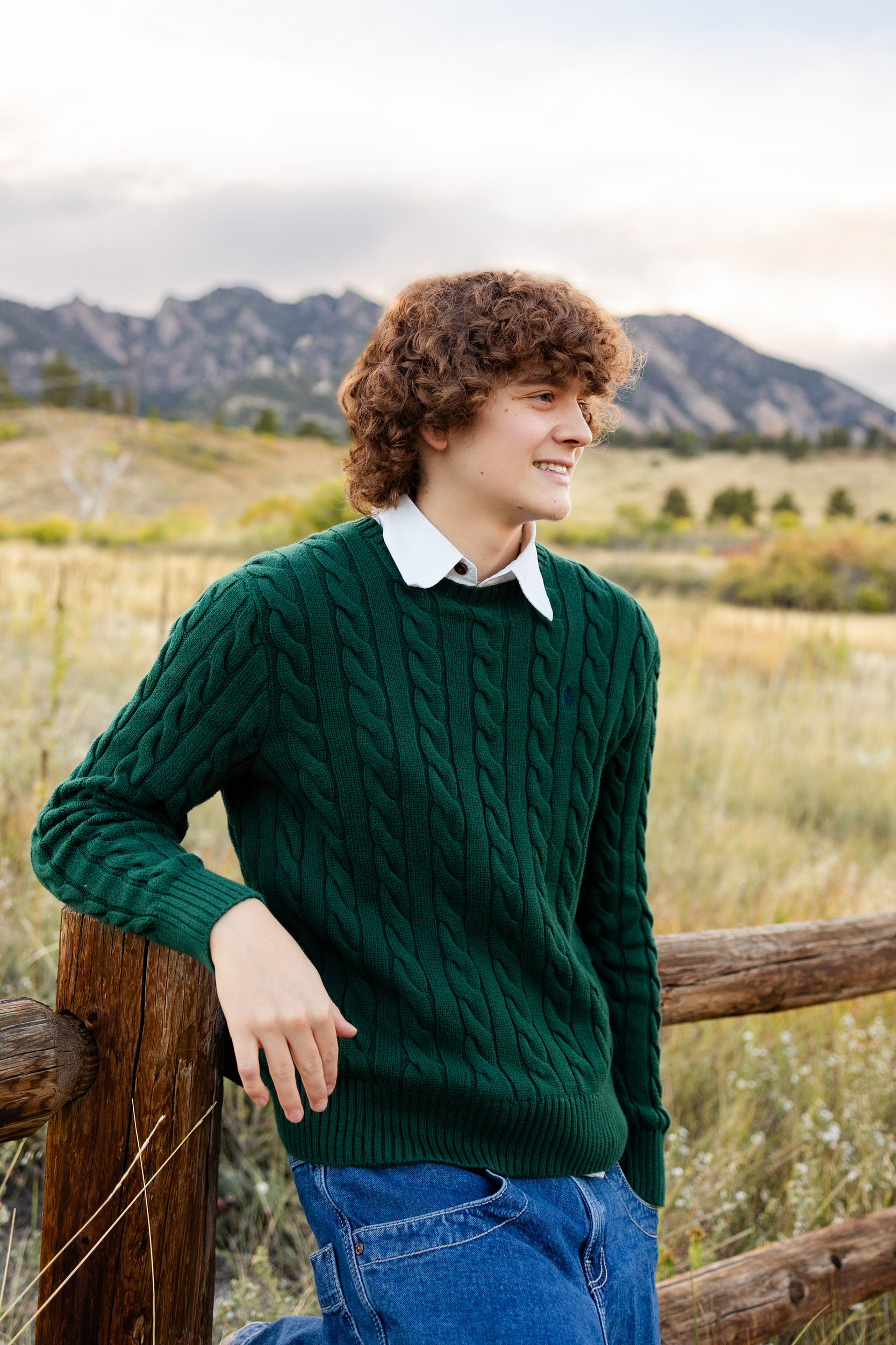 A young man leans against a wooden fence in the mountains.