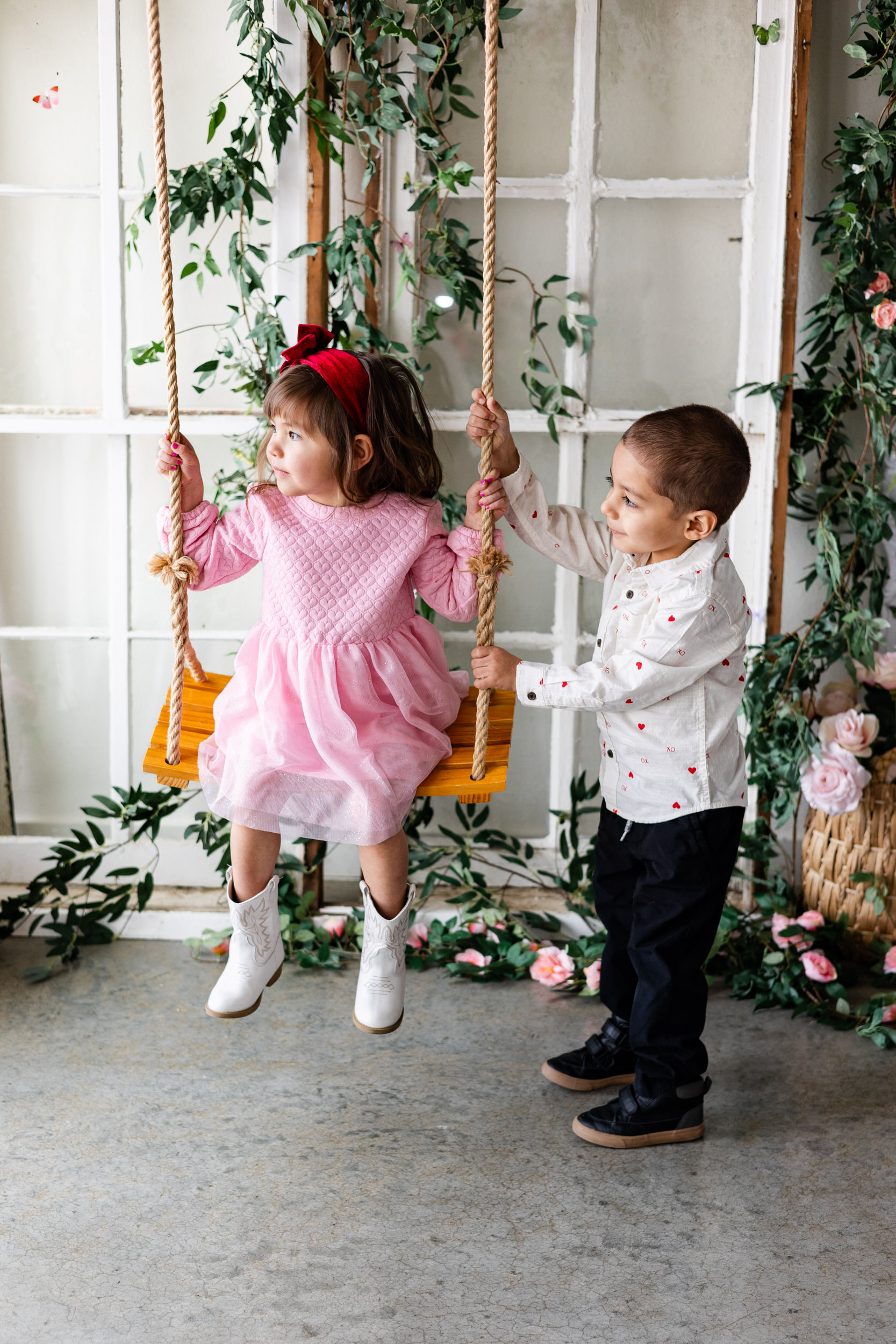 A little boy pushes his younger sister on a swing in front of a wall of flowers.