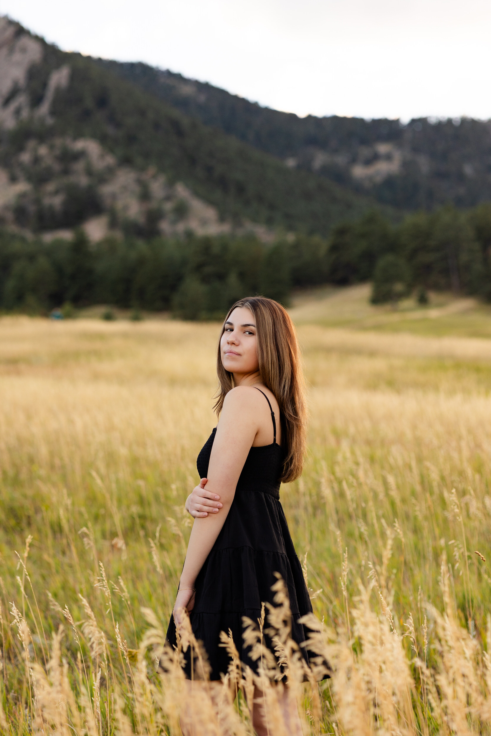 A young woman stands in a field of tall grass in the mountains and looks over her shoulder at the camera.
