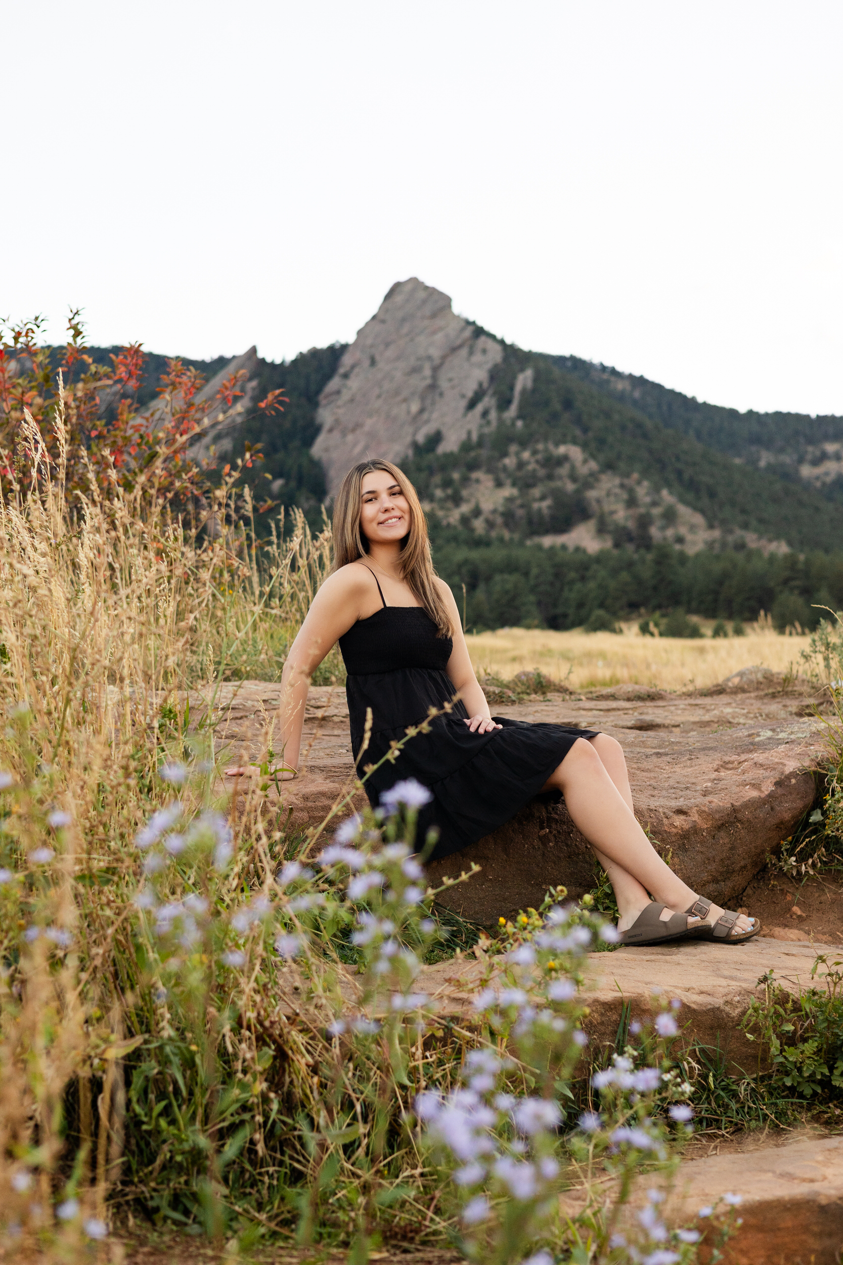 A young woman sits on a rock in Chautauqua Park in Boulder, Colorado and smiles at the camera.