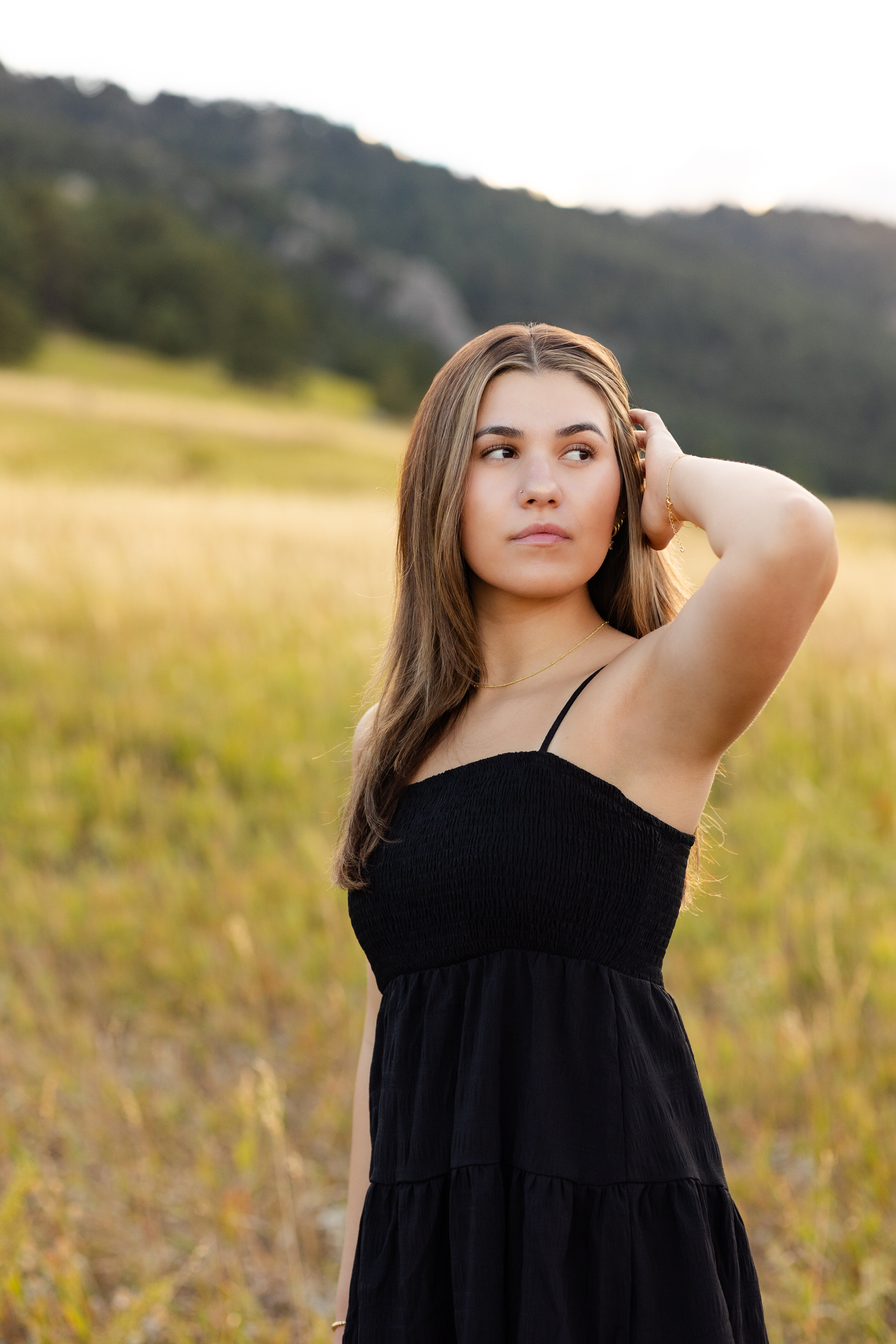 A young woman stands in a field running one hand through her hair and looking over her shoulder off-camera.