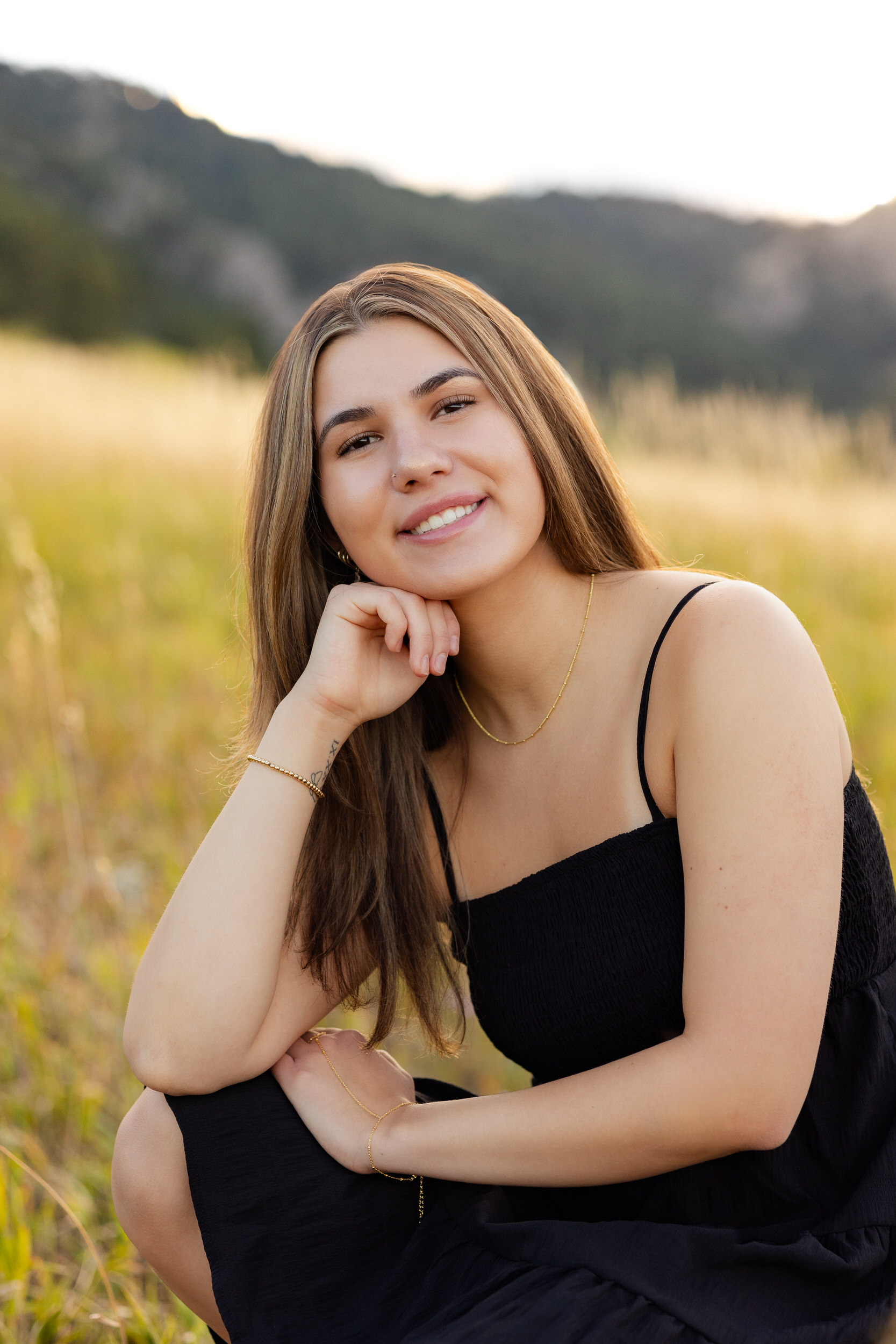 A young woman sits in a field resting her chin on one hand and smiles at the camera.