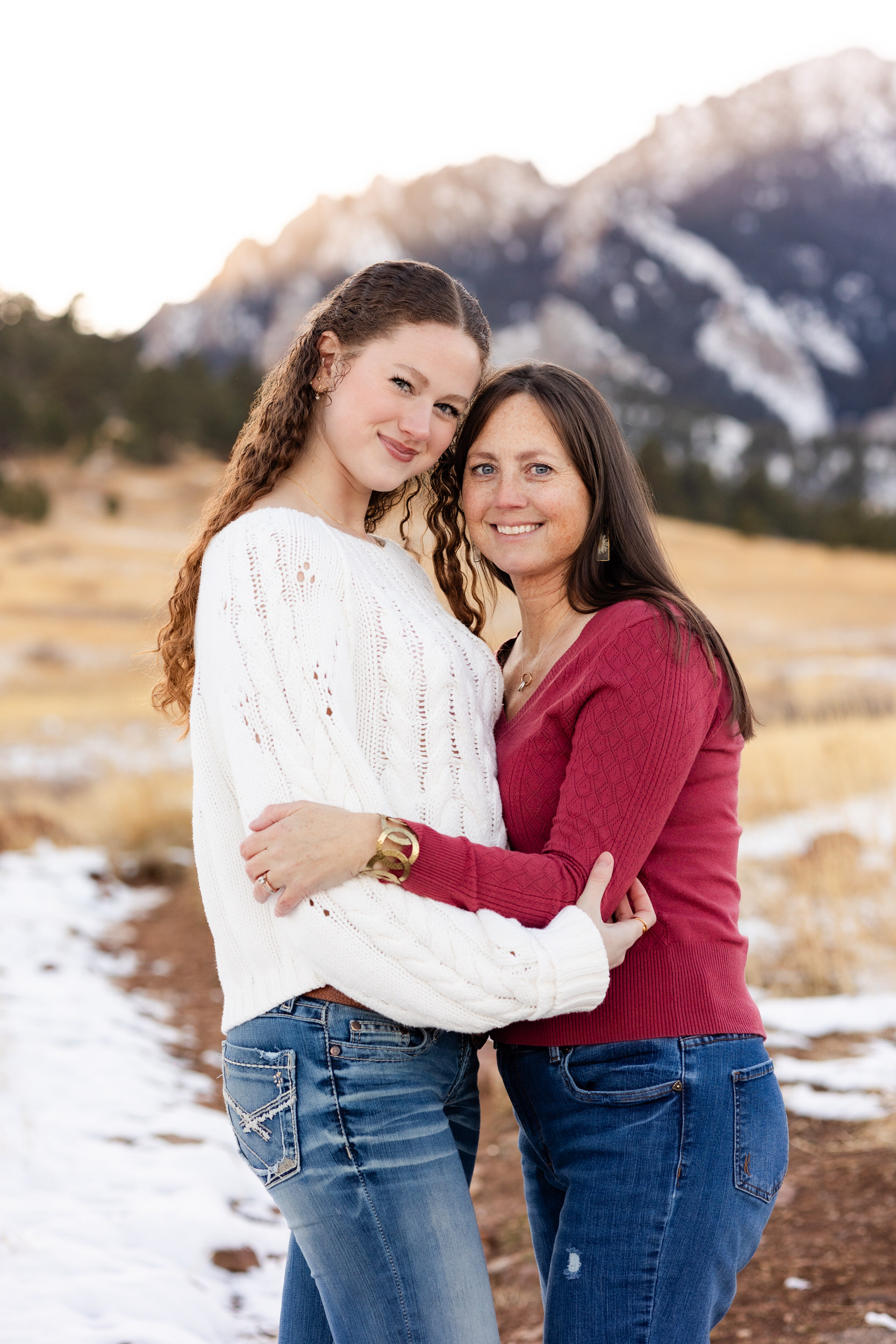 A mom and her adult daughter hug each other and smile at the camera.
